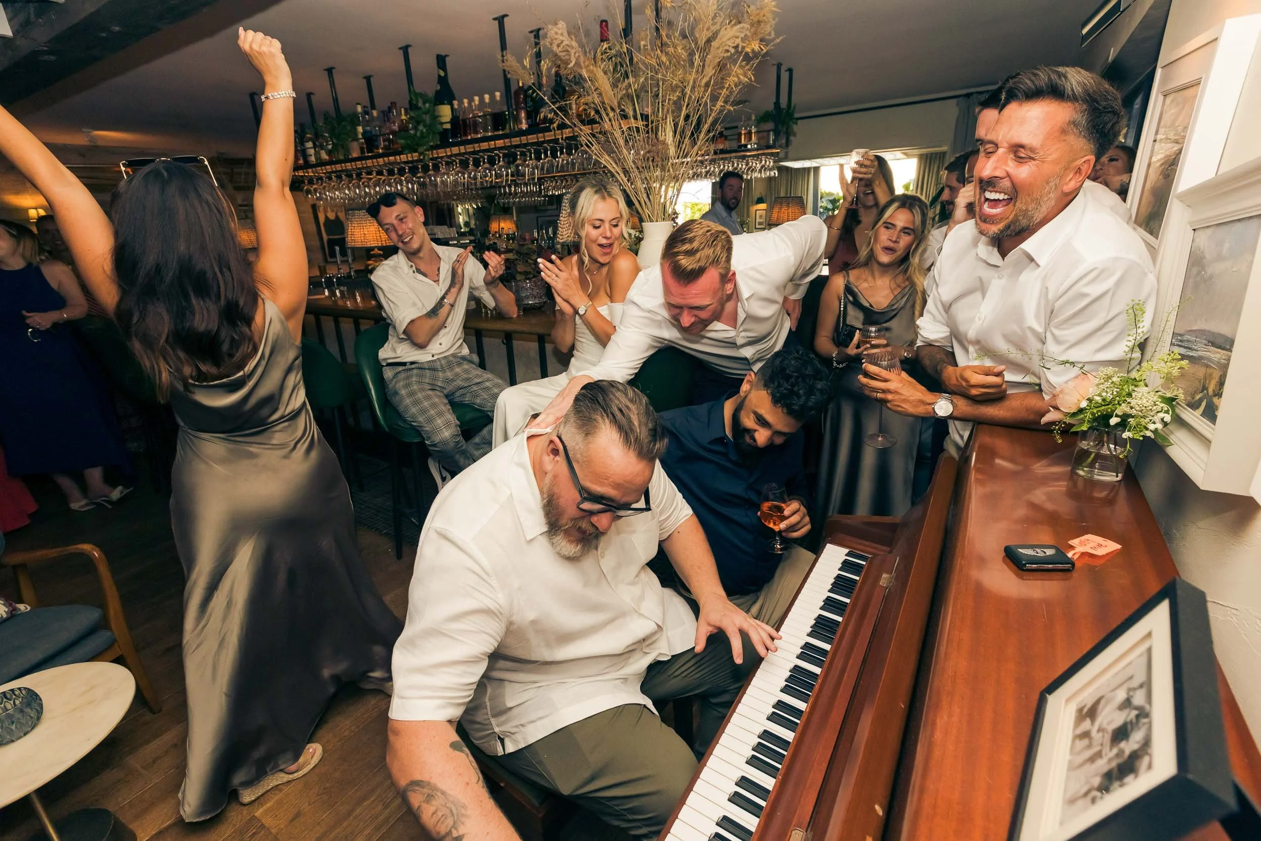 Group of people celebrating around a piano at a lively party in a bar or restaurant. People are dancing, clapping, and laughing, with drinks in hand, creating a joyful atmosphere.