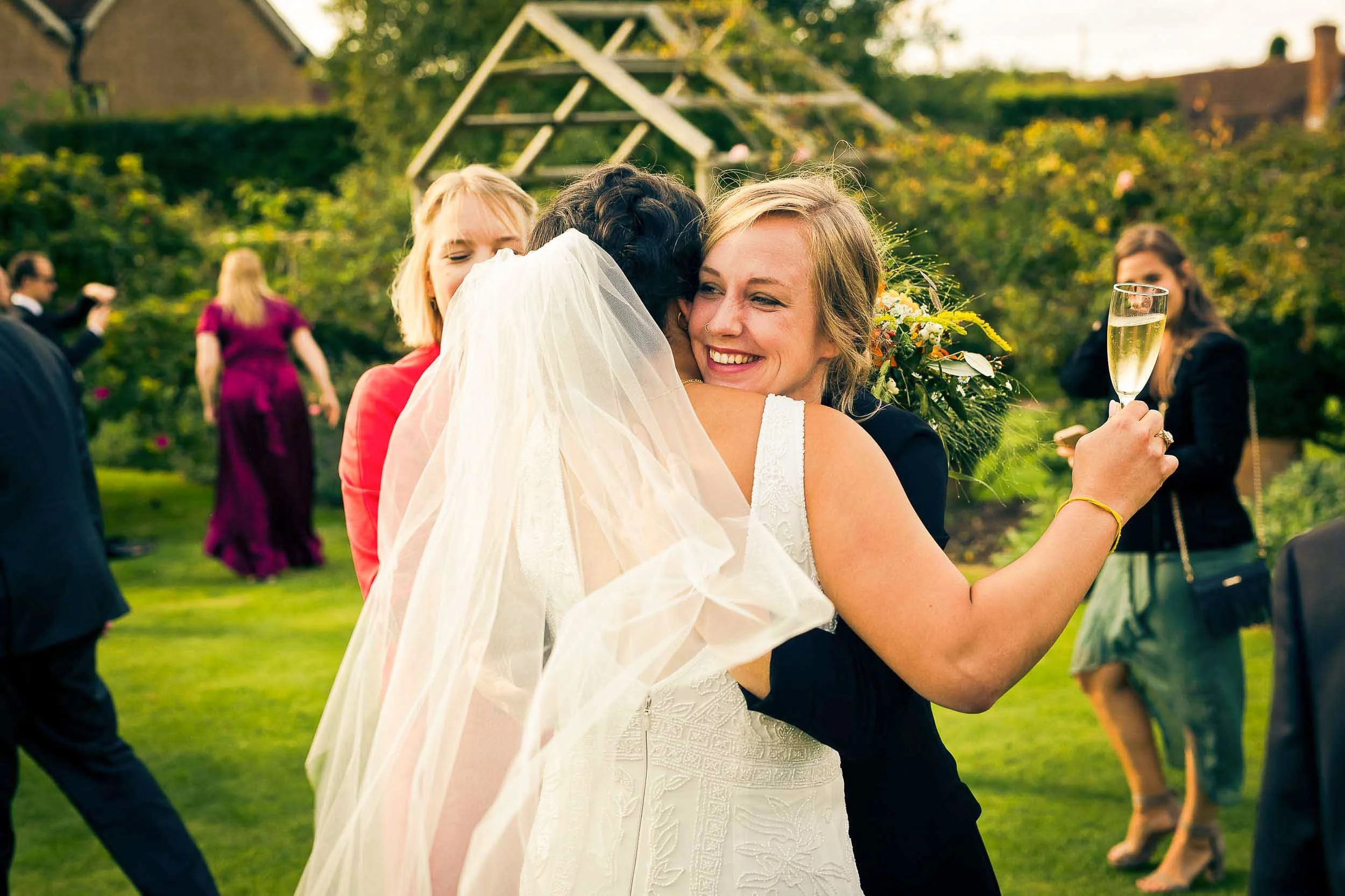 A smiling woman in a wedding dress hugs another woman at an outdoor wedding reception, holding a glass of champagne, with guests and greenery in the background.