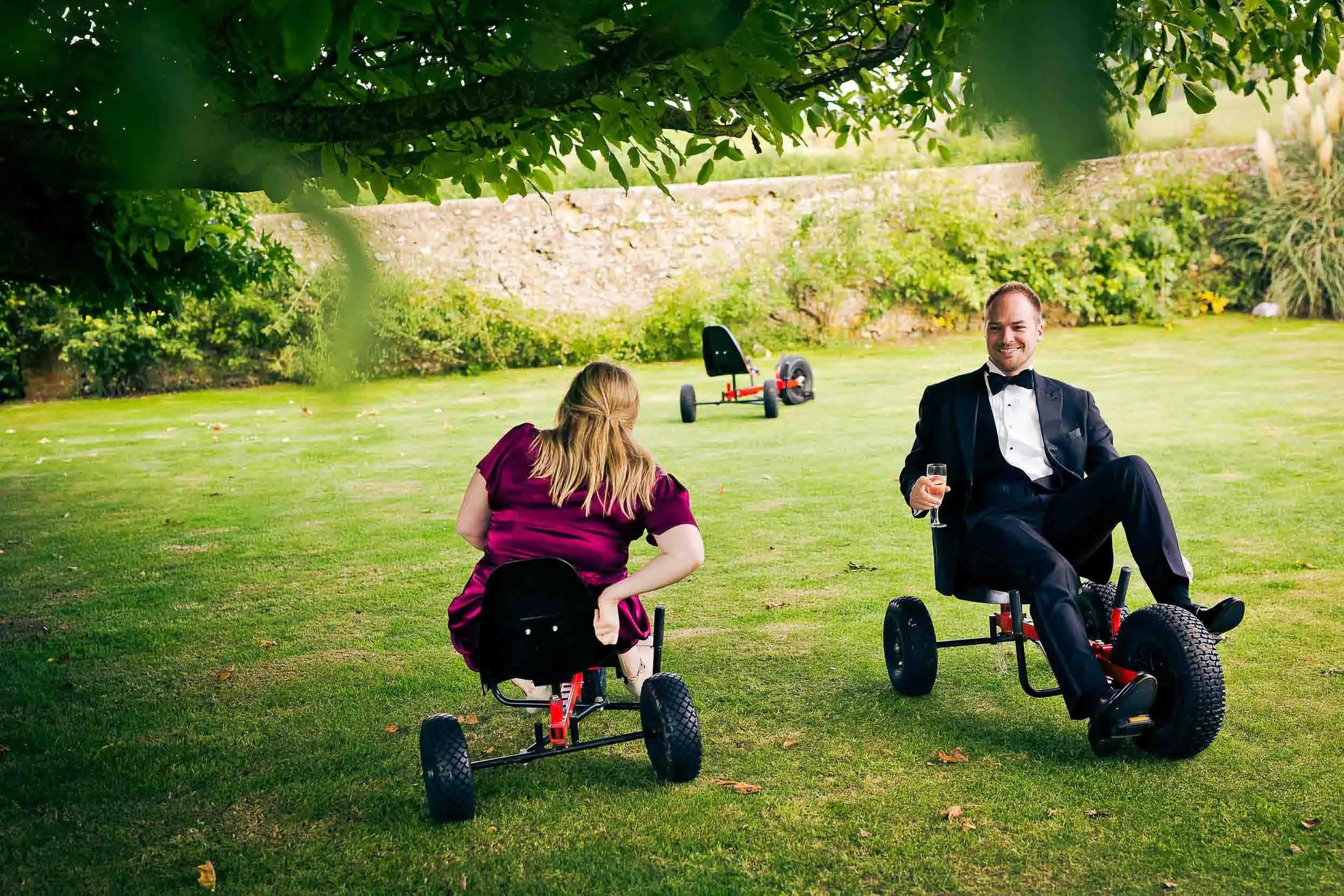 A man in a tuxedo sitting on a mobility vehicle, holding a glass of champagne, smiling in a garden, with a woman sitting on a mobility vehicle nearby, and a dune buggy in the background.