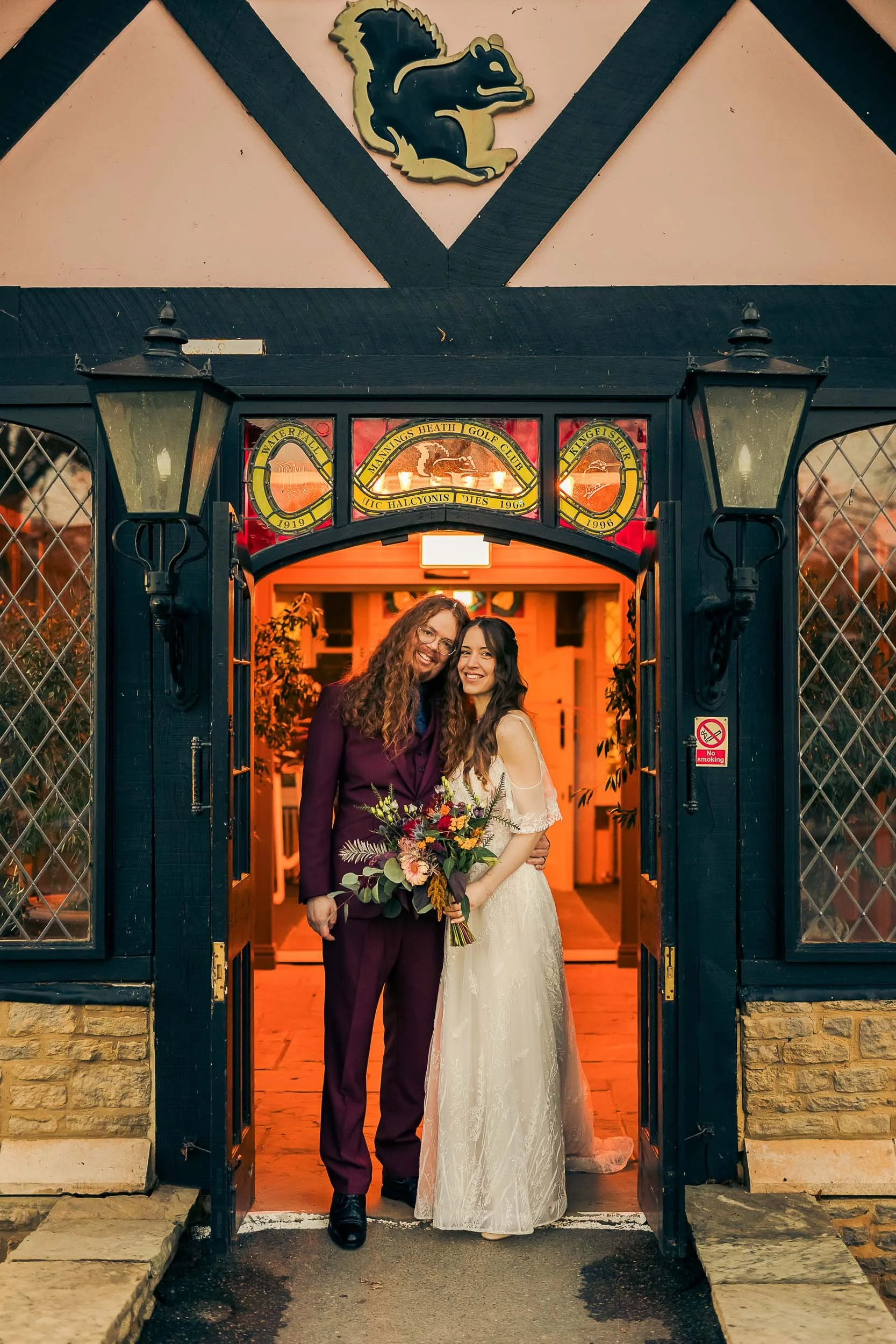 Two women, one in a white wedding dress holding a bouquet of flowers and the other in a dark purple suit, standing close and smiling at the entrance of a building with a Lincolnshire flag above. The entrance has black framing, lanterns, and stained g