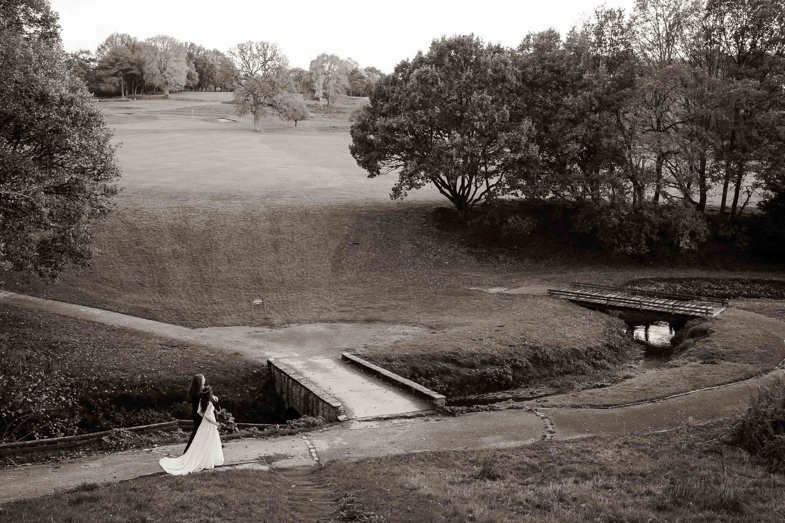 A woman in a wedding dress is walking along a park trail next to a small stream, with trees and open grassy areas in the background.