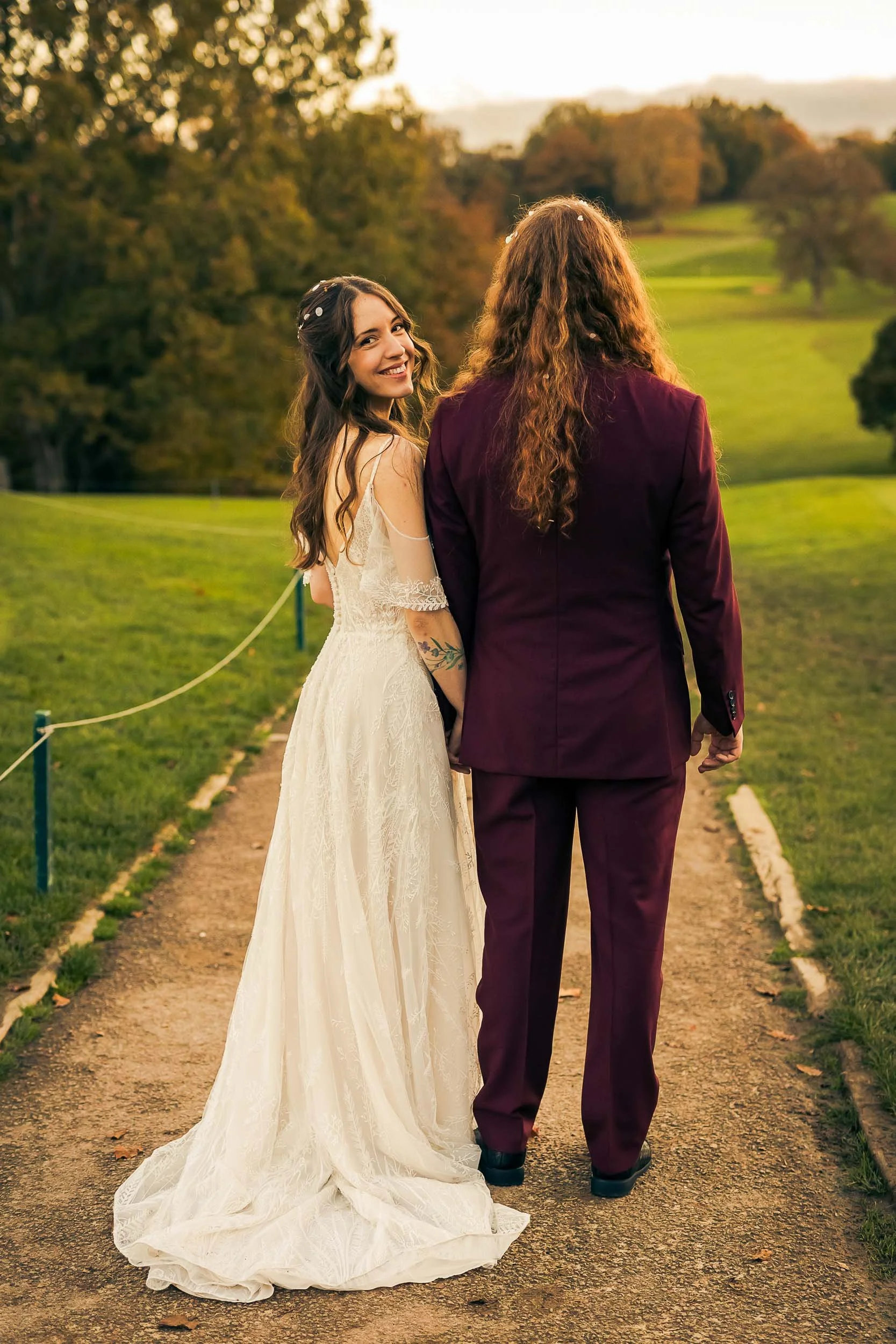 A bride and groom holding hands and walking on a path in a park during sunset, with trees in the background.