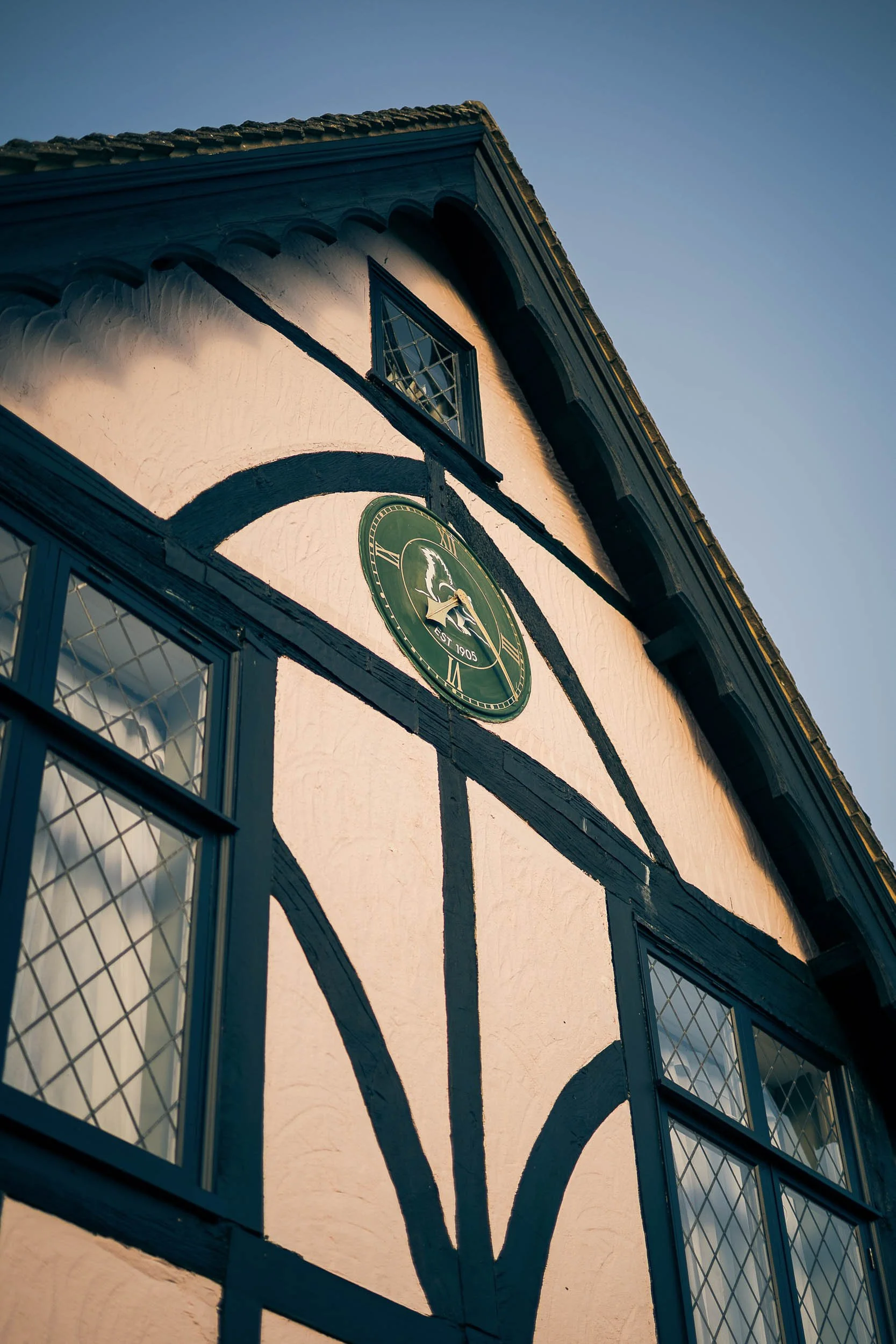 Close-up view of a traditional half-timbered building with black wooden beams and white stucco walls, featuring a round clock with a green face and a decorative house emblem, under a clear blue sky.