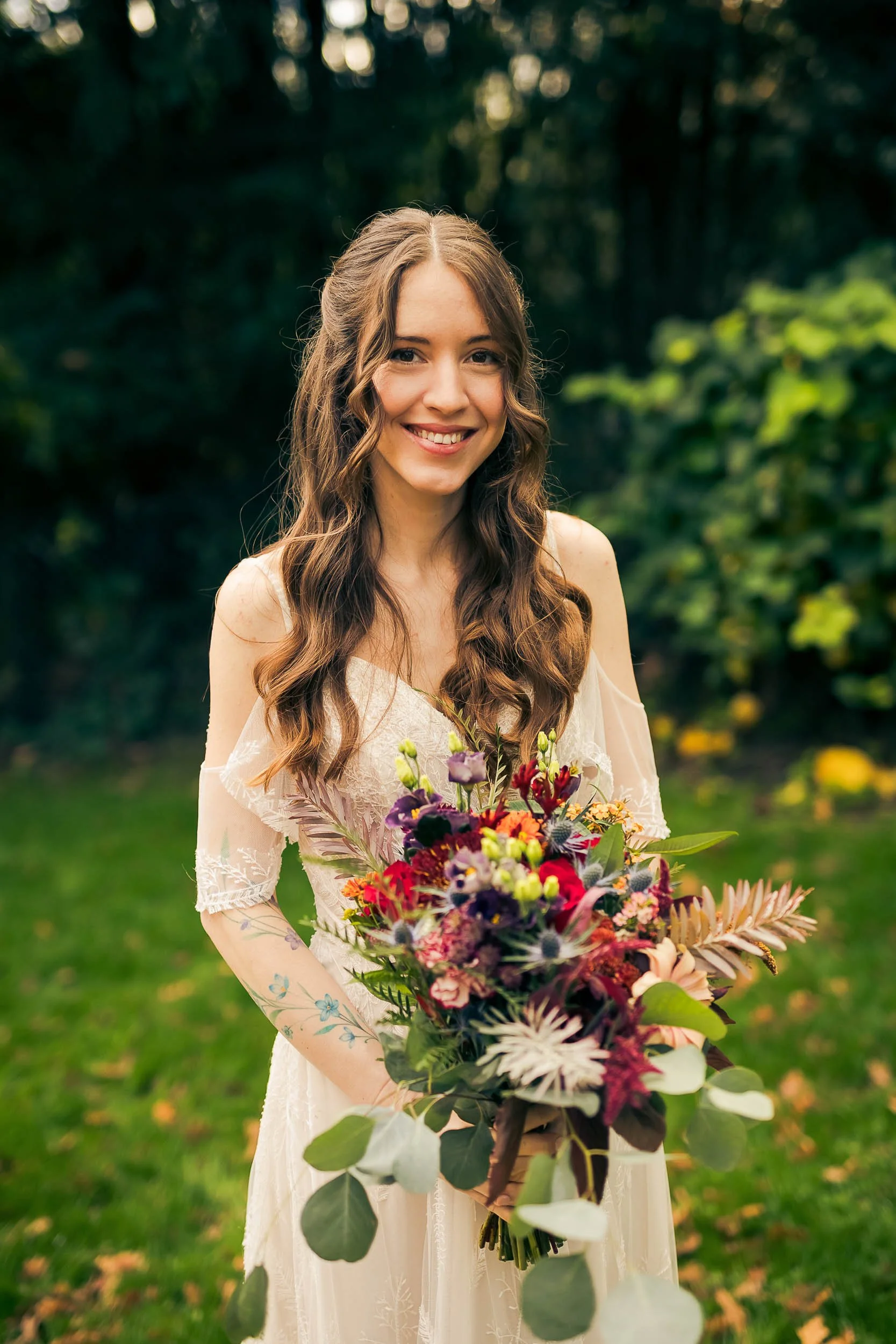 A woman with long wavy brown hair smiling and holding a large colorful bouquet of flowers outdoors in a green, wooded area.