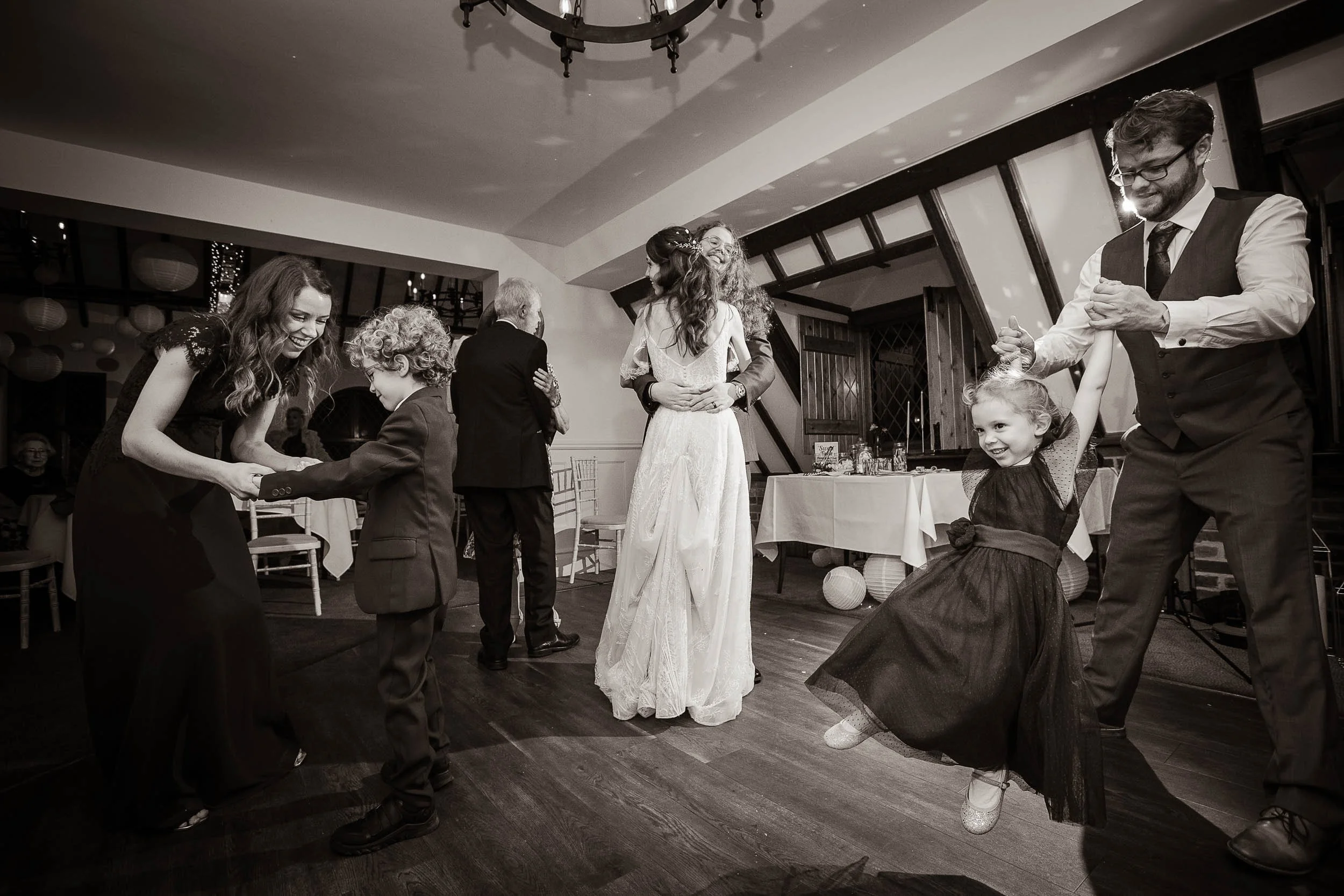 People dancing and enjoying themselves at a wedding reception, with a woman in a wedding dress in the center hugging someone, and children and adults dancing around.