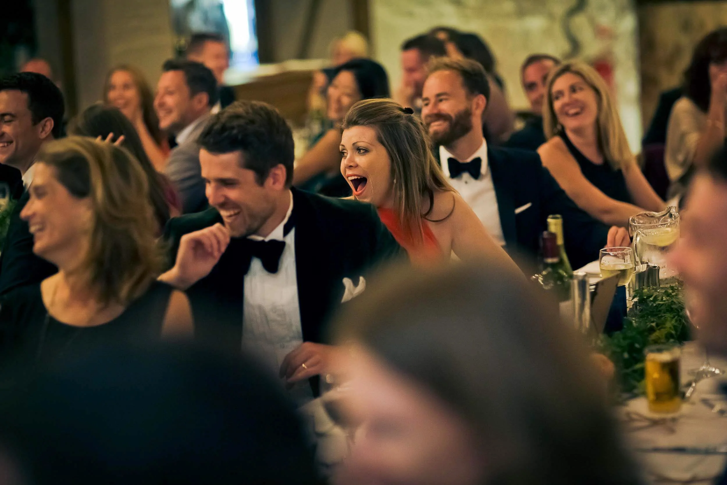 Guests at a formal event, laughing and enjoying themselves, with men in tuxedos and women in dresses, seated at a banquet table with drinks.