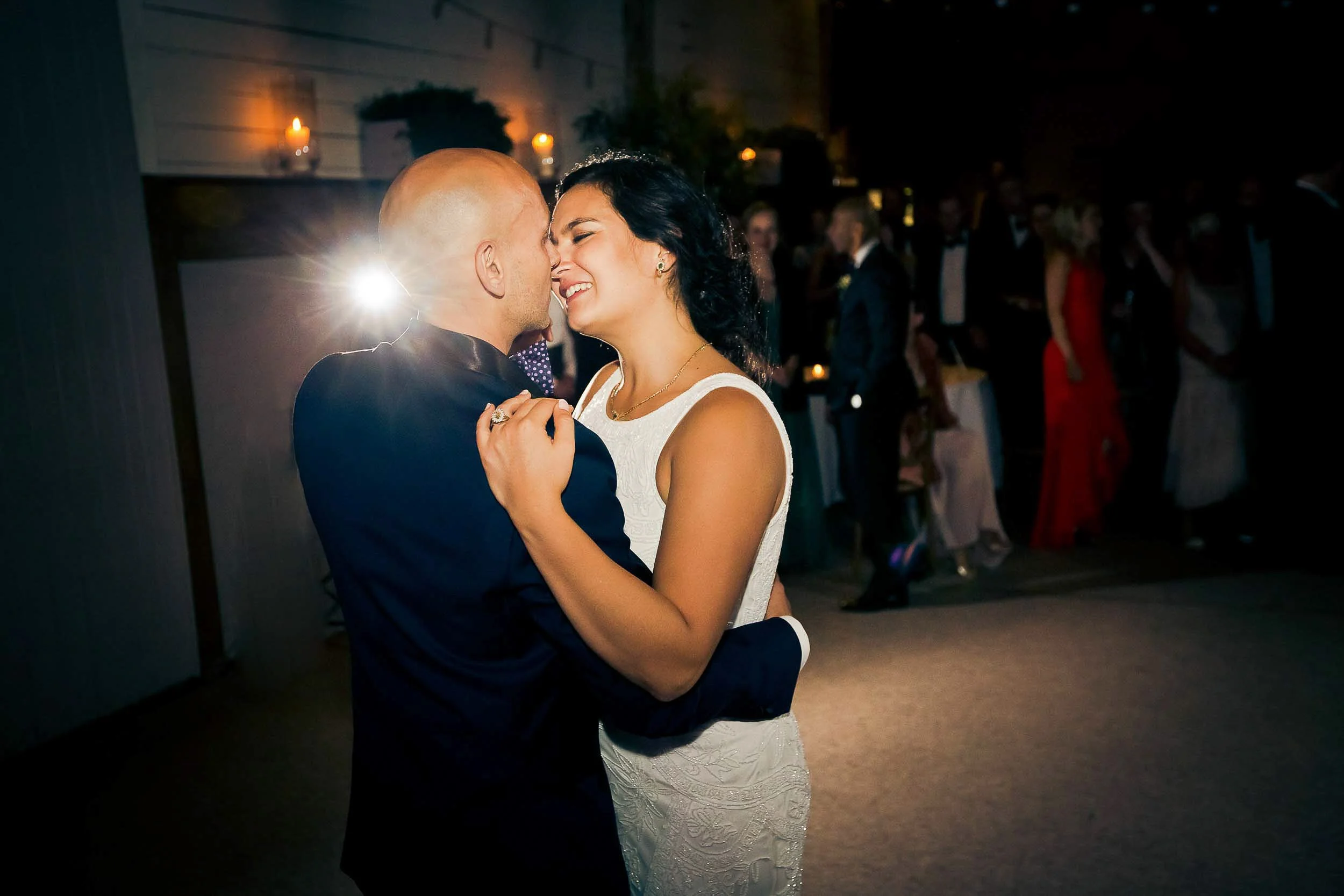 A newlywed bride and groom share a dance, smiling with their faces close, in a dimly lit wedding reception hall with guests in the background.