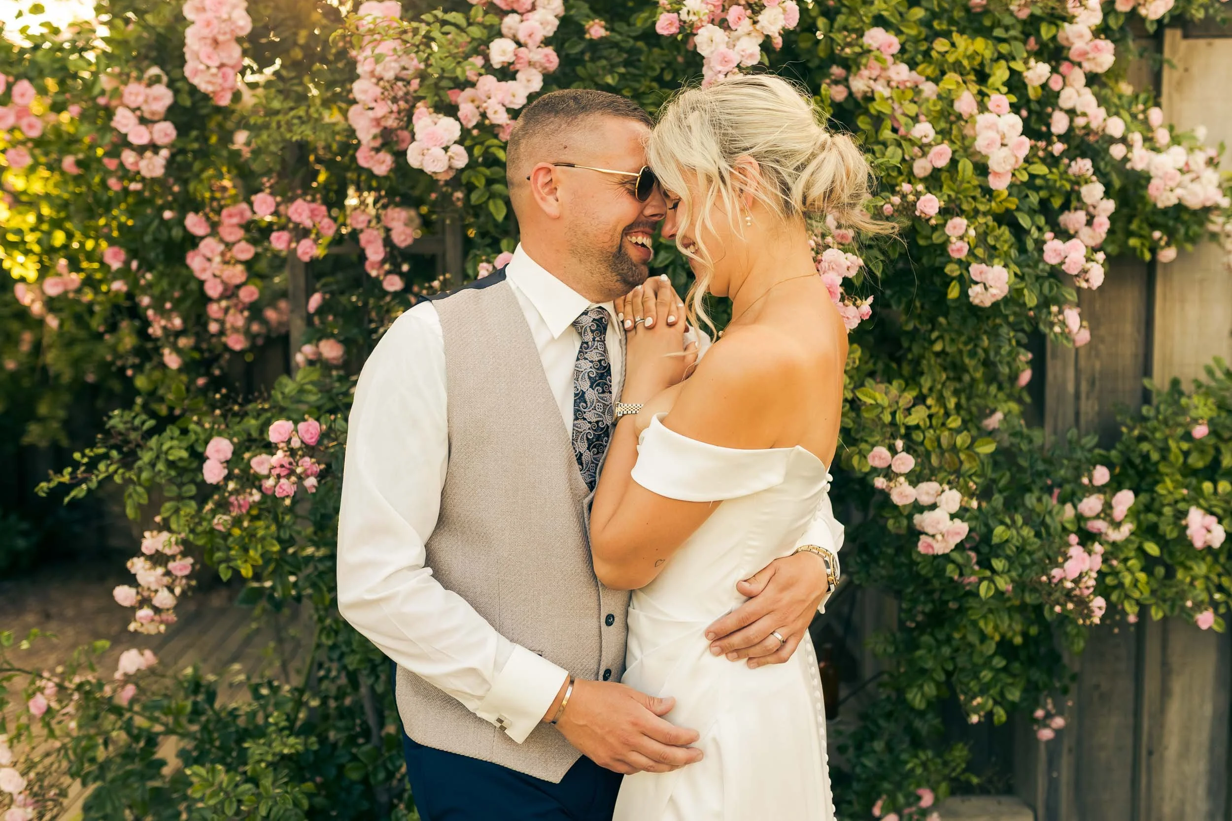 A happy couple, a man and woman, close together and smiling, outdoor setting with blooming pink flowers in background.