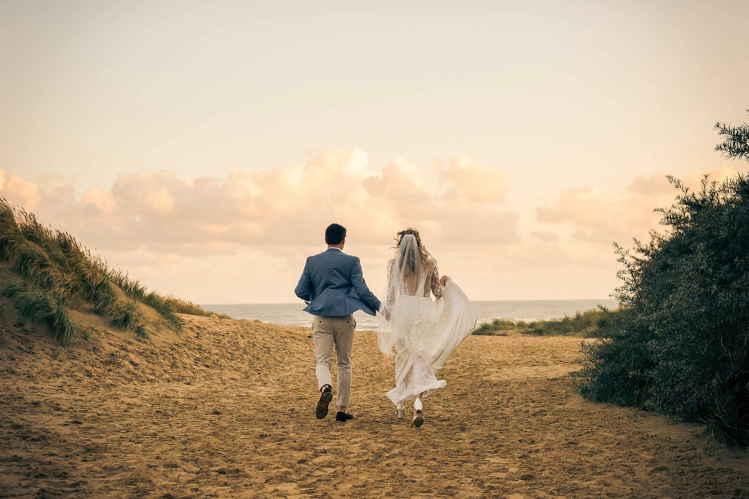 A newlywed couple walking hand in hand on a sandy beach during sunset, with the ocean and clouds in the background.