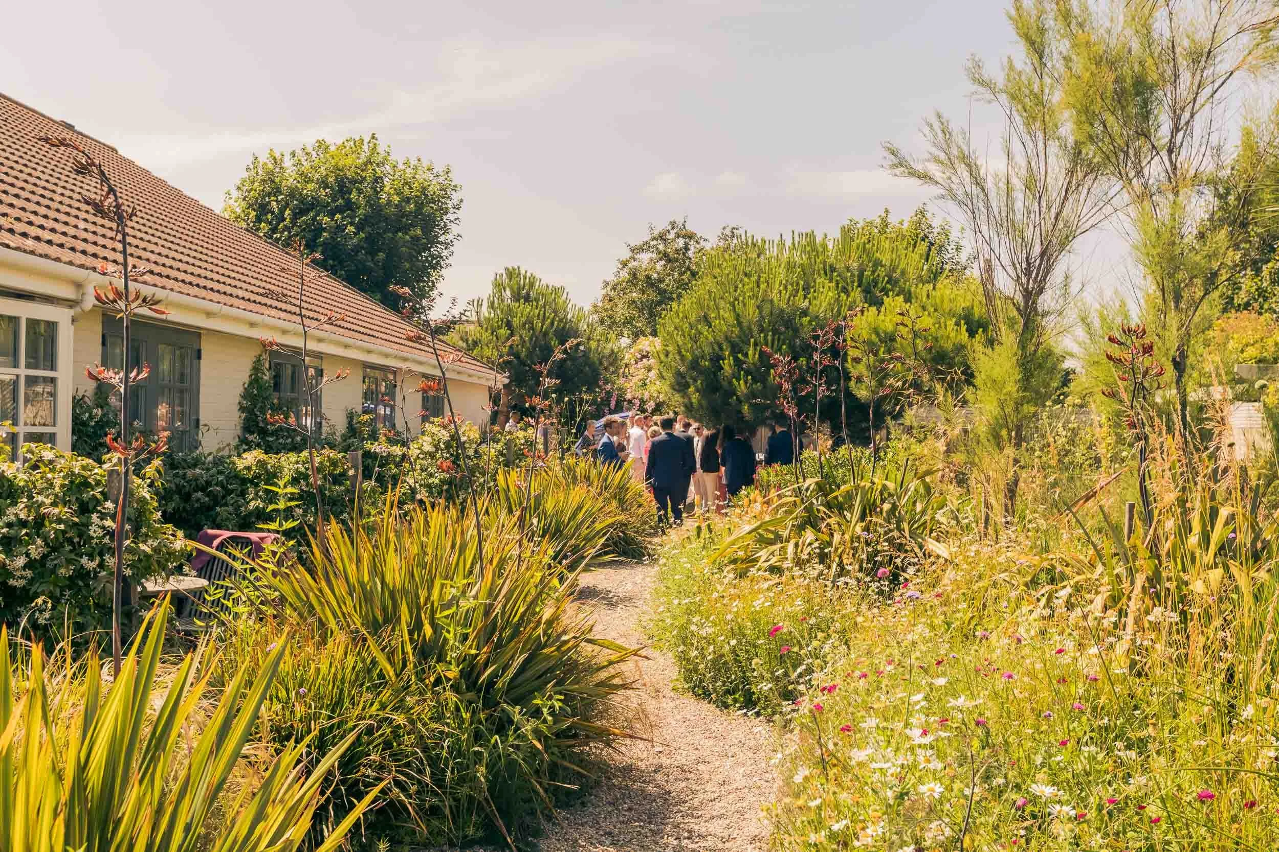 Group of people walking along a garden pathway next to a house with a tiled roof, surrounded by tall trees and various flowering plants on a sunny day.