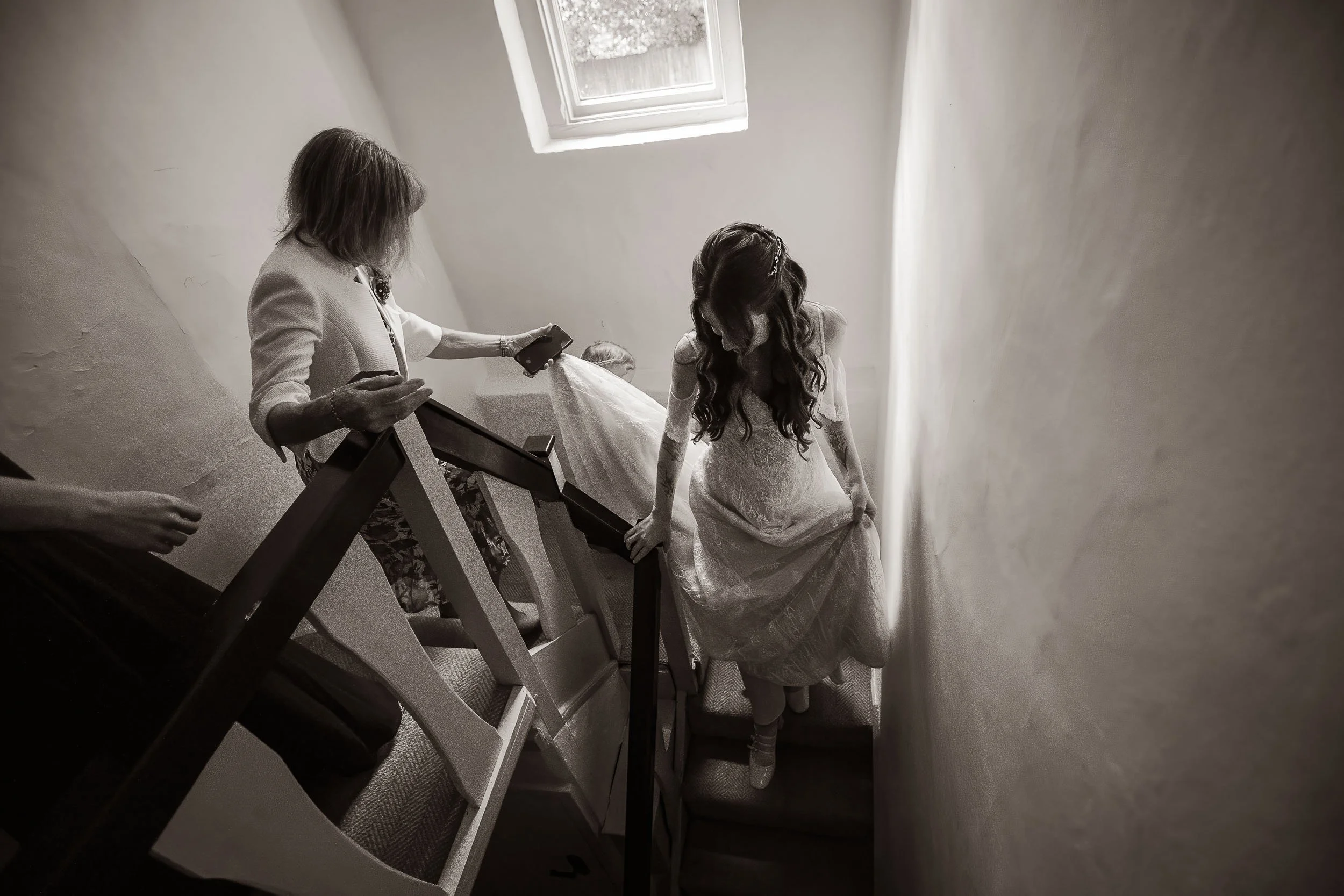 A bride in a white wedding dress standing on a staircase at the top step, while two women, one holding a phone, assist her. The scene is in a narrow stairwell with a small window on the ceiling allowing natural light in.