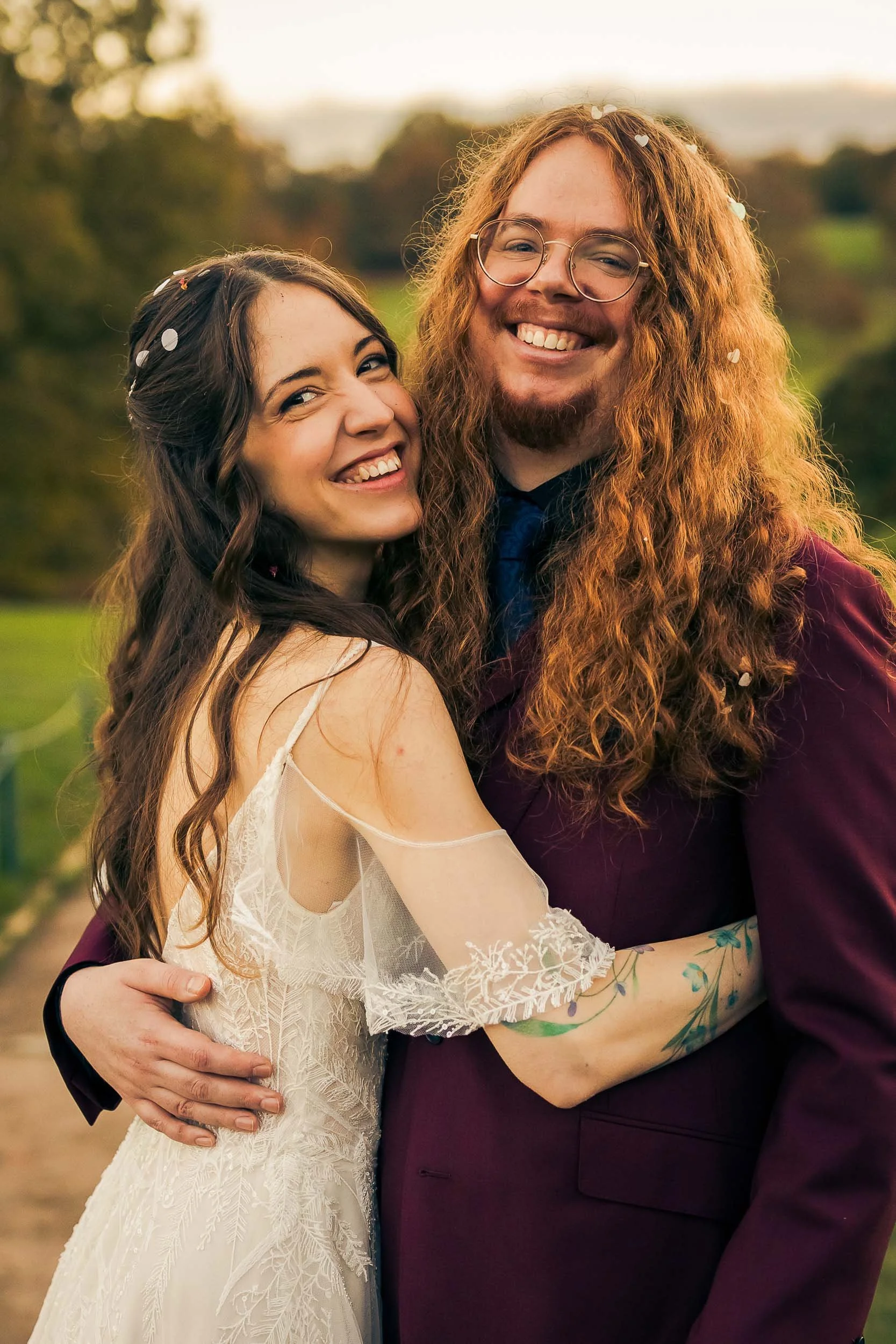 A happy couple hugging outdoors during sunset, woman in a white wedding dress and man in a dark suit with glasses and long curly hair, surrounded by falling confetti.