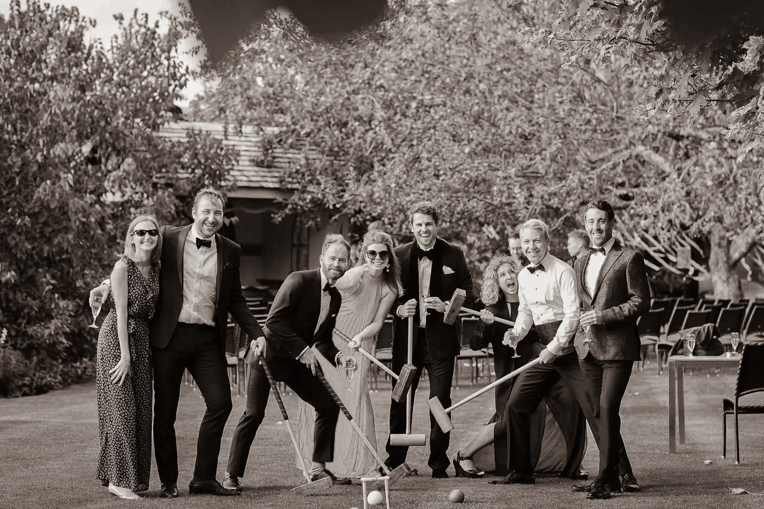 Group of people in formal attire playing croquet outdoors, smiling and laughing, in a garden with trees and a cottage in the background, all captured in black and white.