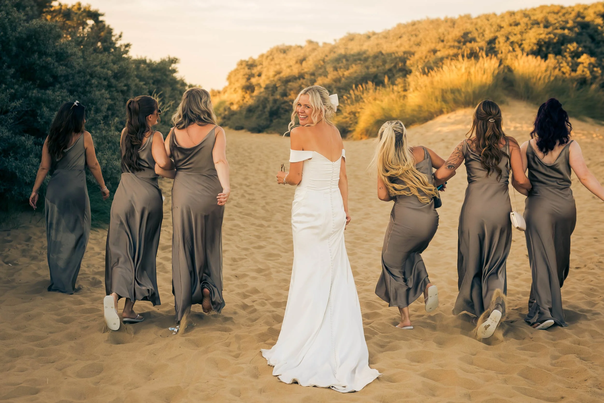 A bride in a white wedding dress walking on a sandy path with seven bridesmaids in matching gray dresses, all smiling and holding hands in an outdoor setting with trees and a hill in the background.