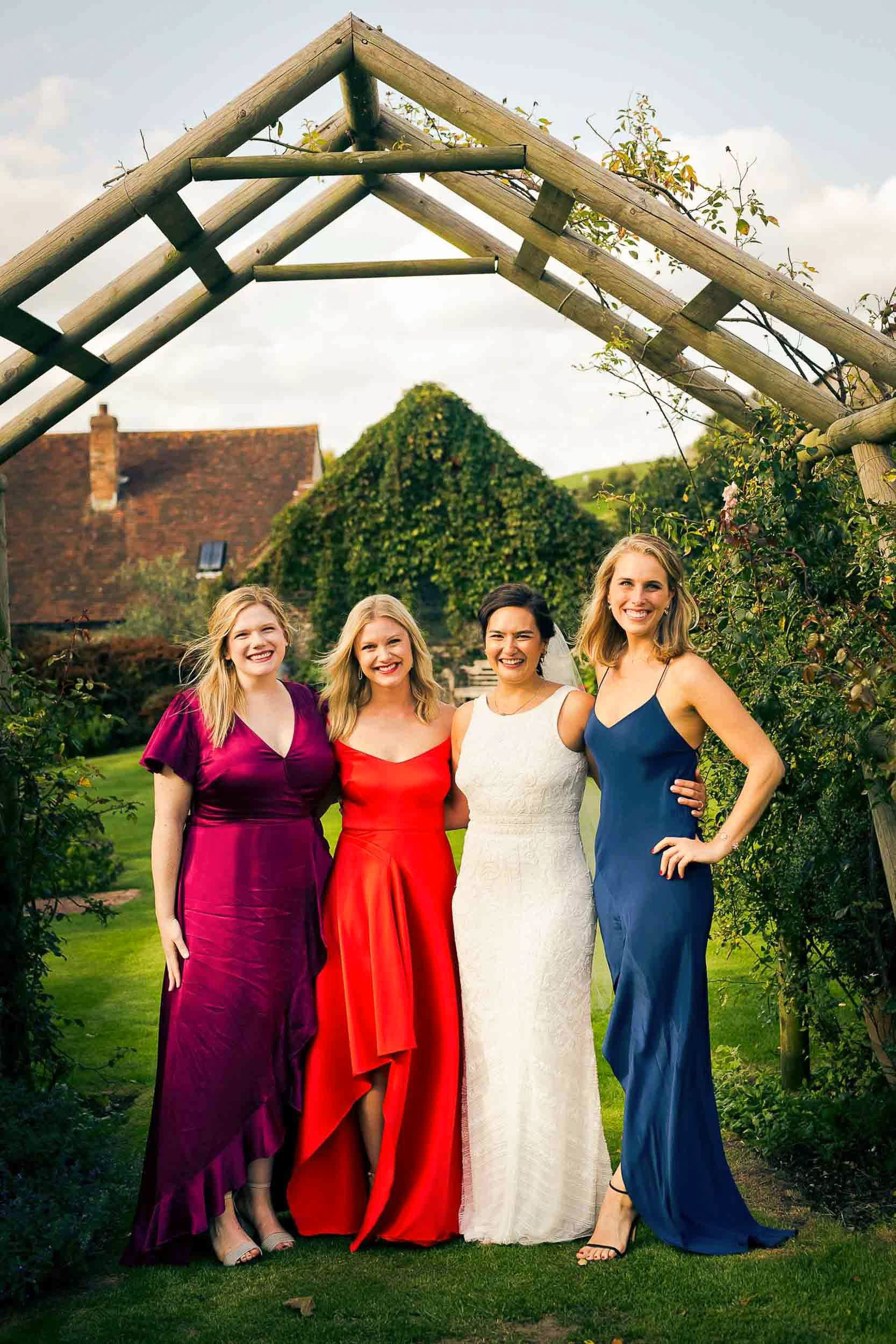 Four women in colorful dresses standing together outdoors beneath a wooden arch, with a garden and house in the background, smiling at the camera.