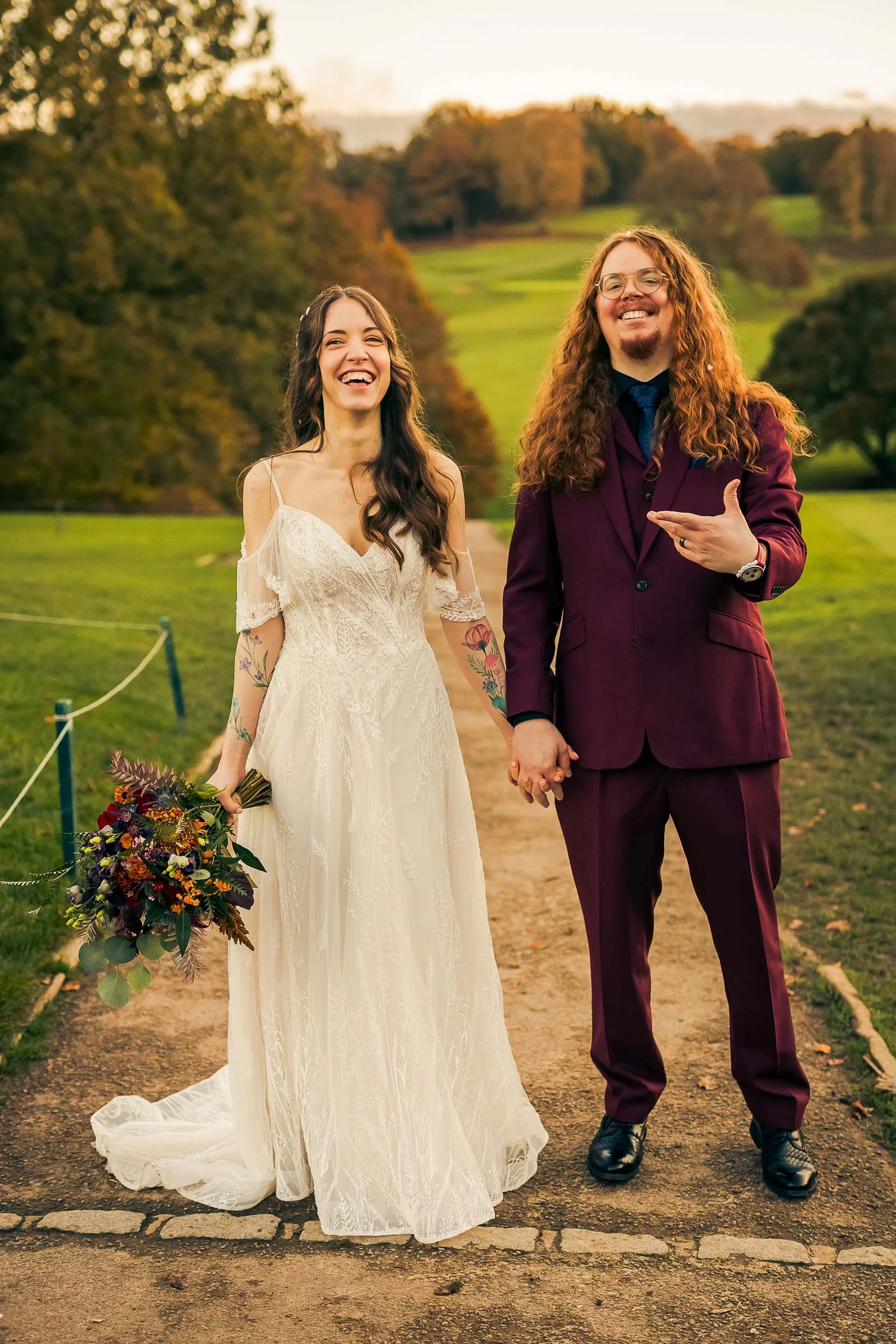 A happy couple in wedding attire holding hands on a scenic outdoor path with trees and a green landscape in the background during sunset.