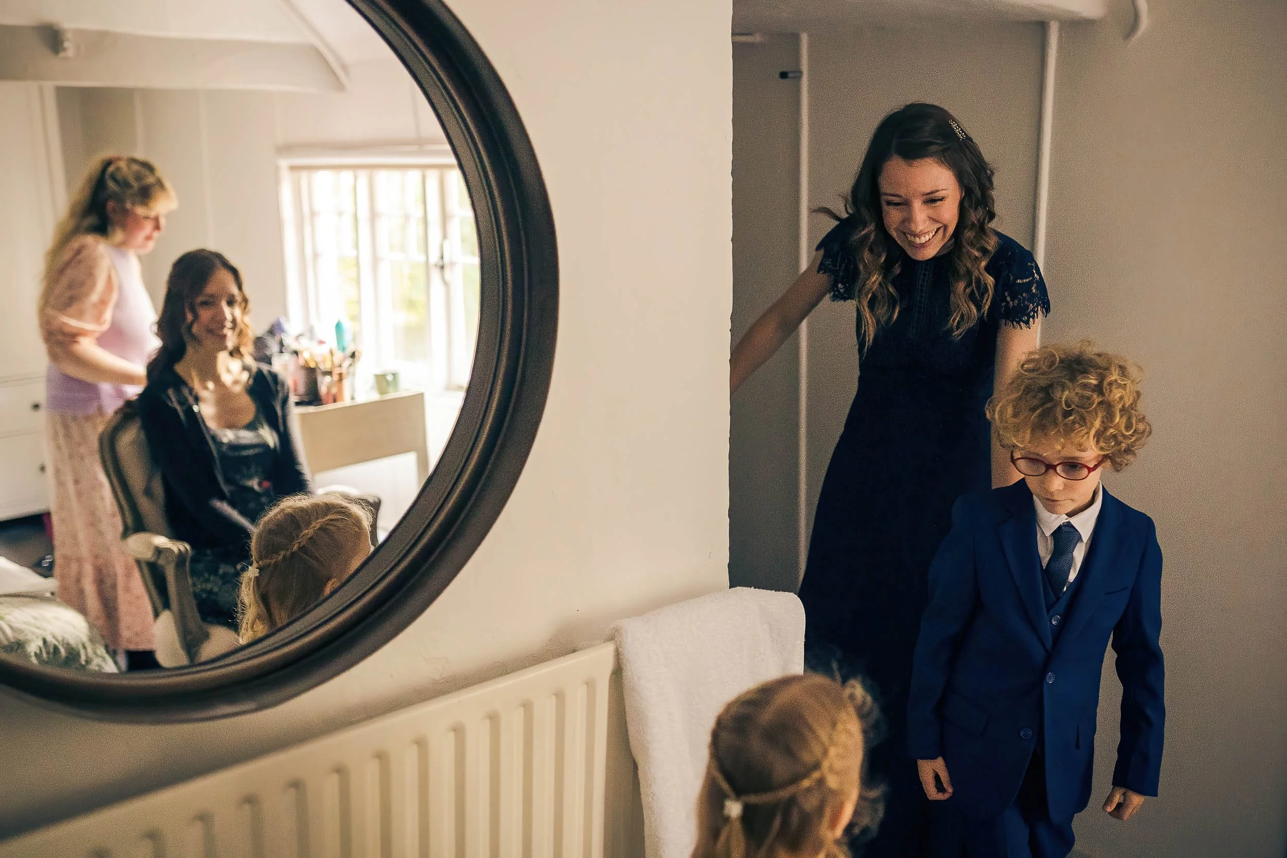 A woman and a young boy in a blue suit and glasses are smiling as they enter a room, while a girl with braided hair watches them from below. A mirror in the room reflects two women, one with curly hair and a floral dress and another with long hair an
