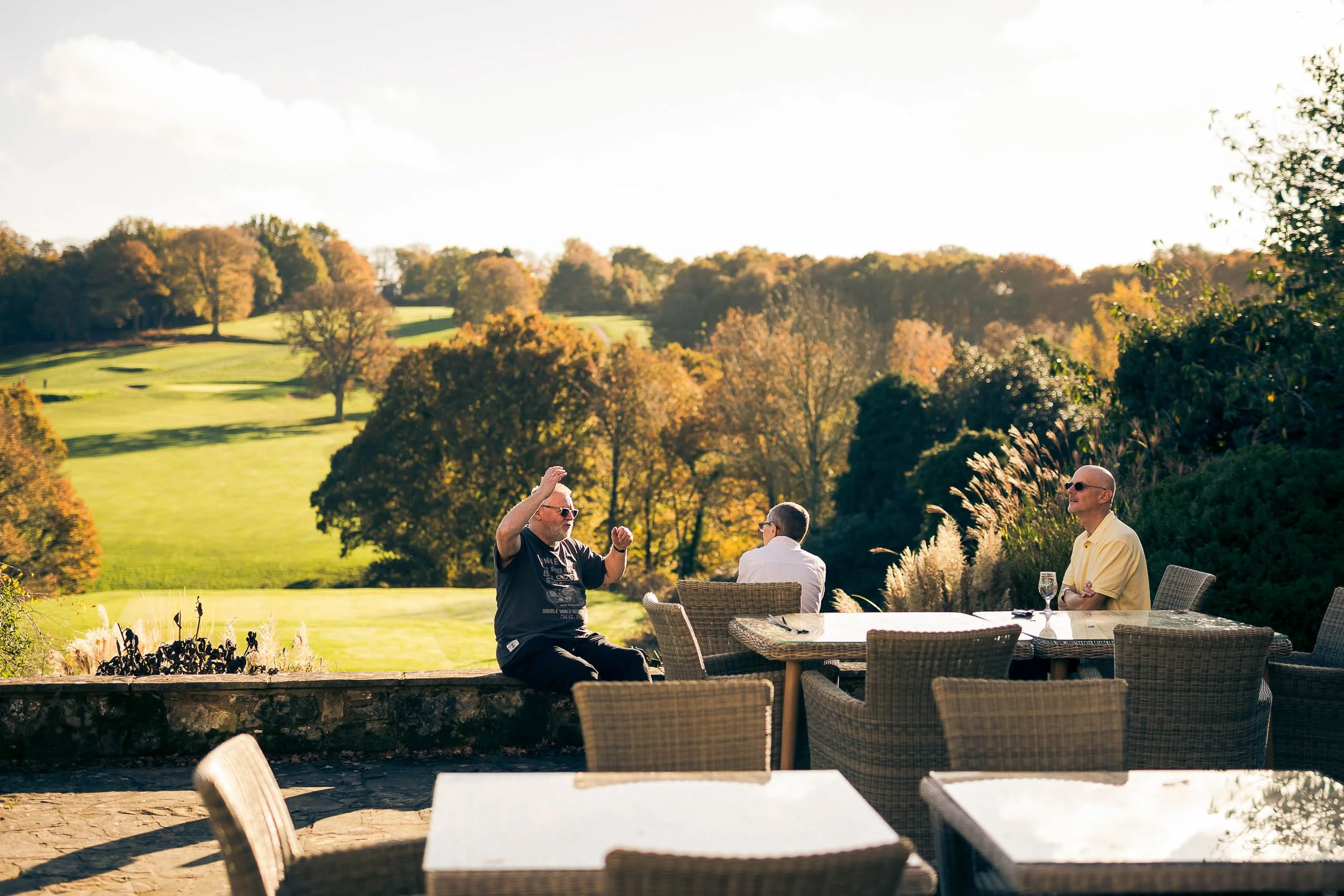 Three men sit and stand at an outdoor table on a patio overlooking a lush golf course and trees in autumn.