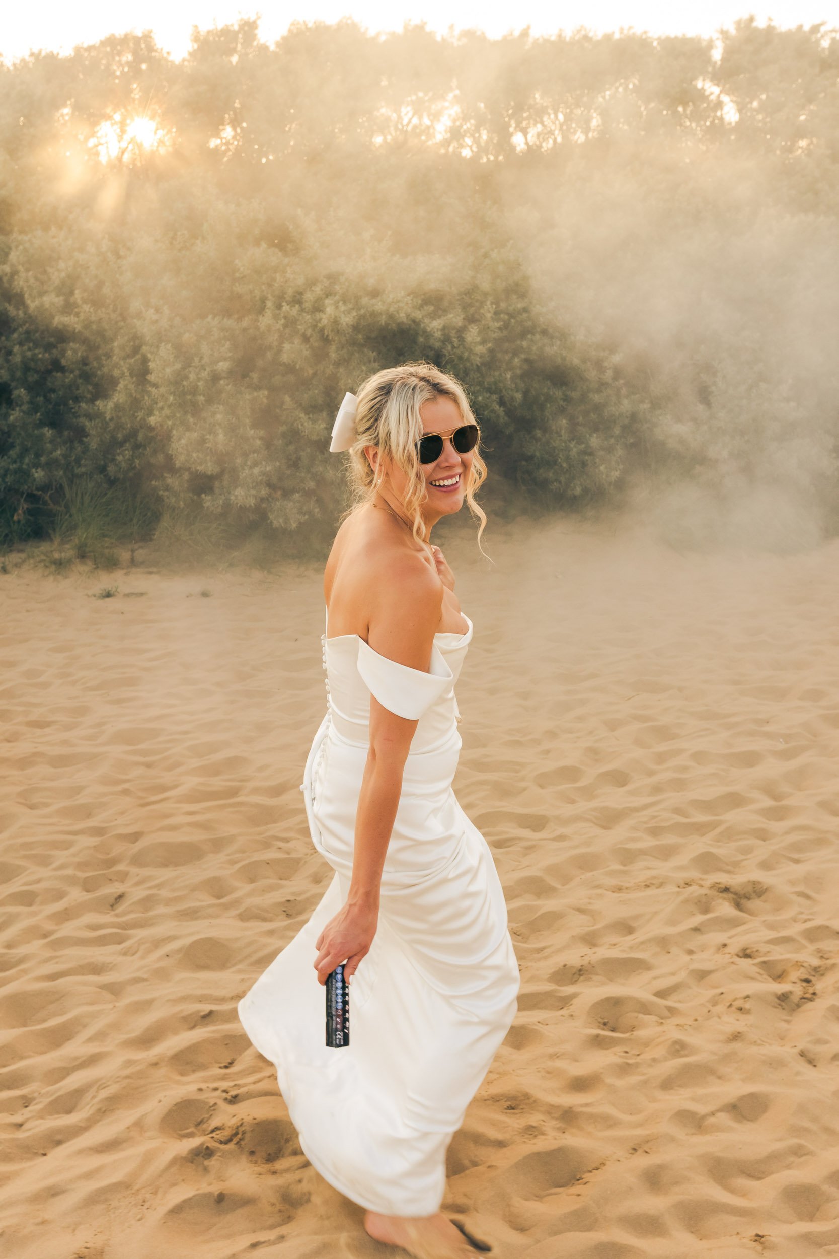 Woman in a white off-shoulder dress and black sunglasses smiling on sandy beach during sunset.