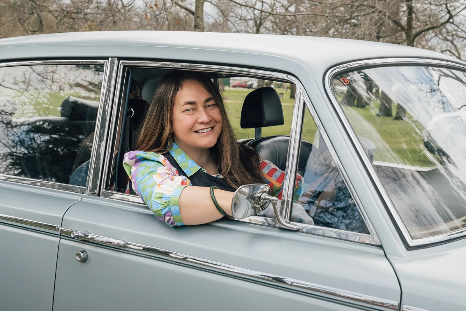 Chauffeuse (female chauffeur) smiling and sitting in the driver's seat of a vintage silver shadow Rolls Royce car, wearing a colorful checkered shirt, with a park and trees in the background.