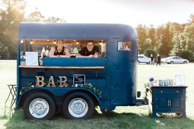A blue mobile bar converted from a trailer with two people inside, parked on a grassy area. The trailer has decorative letters spelling "BAR" on the side. A small table with drinks and a sign is set up next to the trailer.