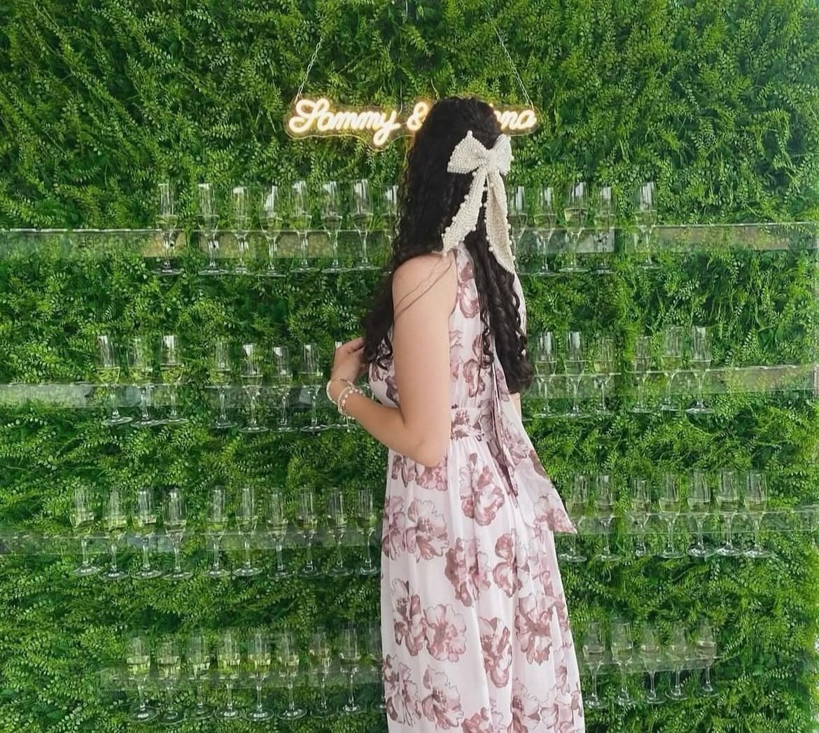 Woman in floral dress facing a greenery wall with champagne glasses