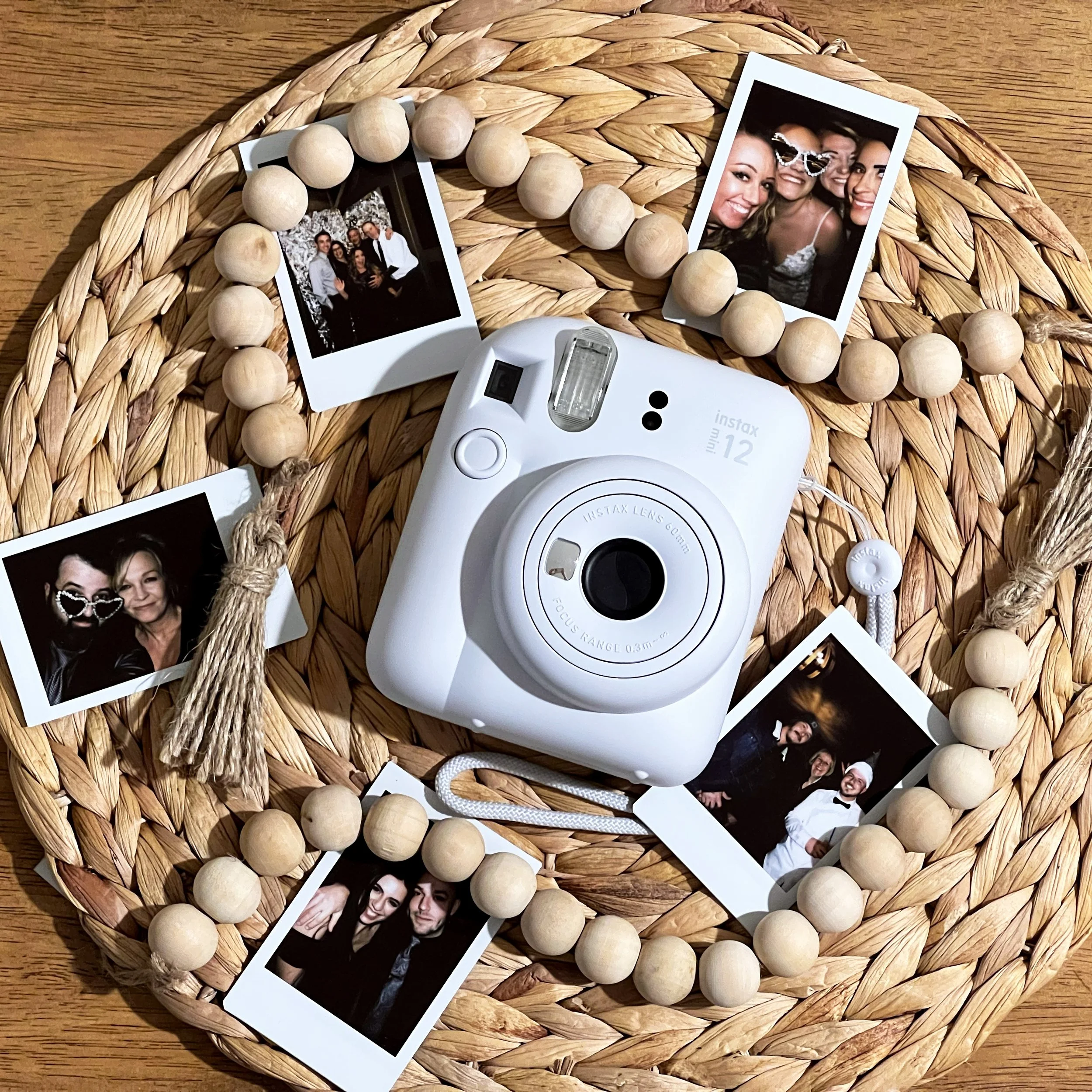 A white Instax Mini 12 camera surrounded by several polaroid photos on a woven placemat, with wooden bead garland accenting the arrangement.