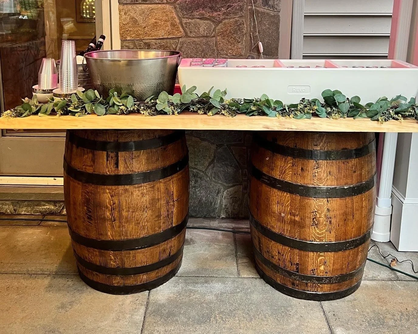 A rustic beverage station setup using two wooden barrels as supports for a wooden plank table. The table is adorned with greenery and includes a metal ice bucket, plastic cups, and a large white cooler. The background features a stone wall and siding.