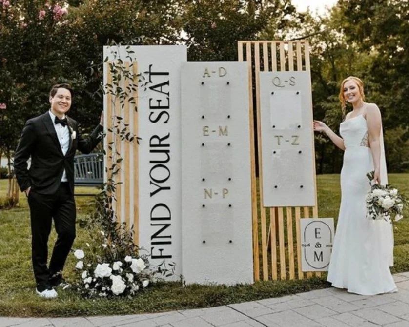 A bride and groom stand on either side of a wedding seating chart display labeled 'Find Your Seat.' The groom is in a tuxedo, and the bride wears a white dress holding a bouquet. The chart is organized alphabetically and decorated with greenery and flowers.