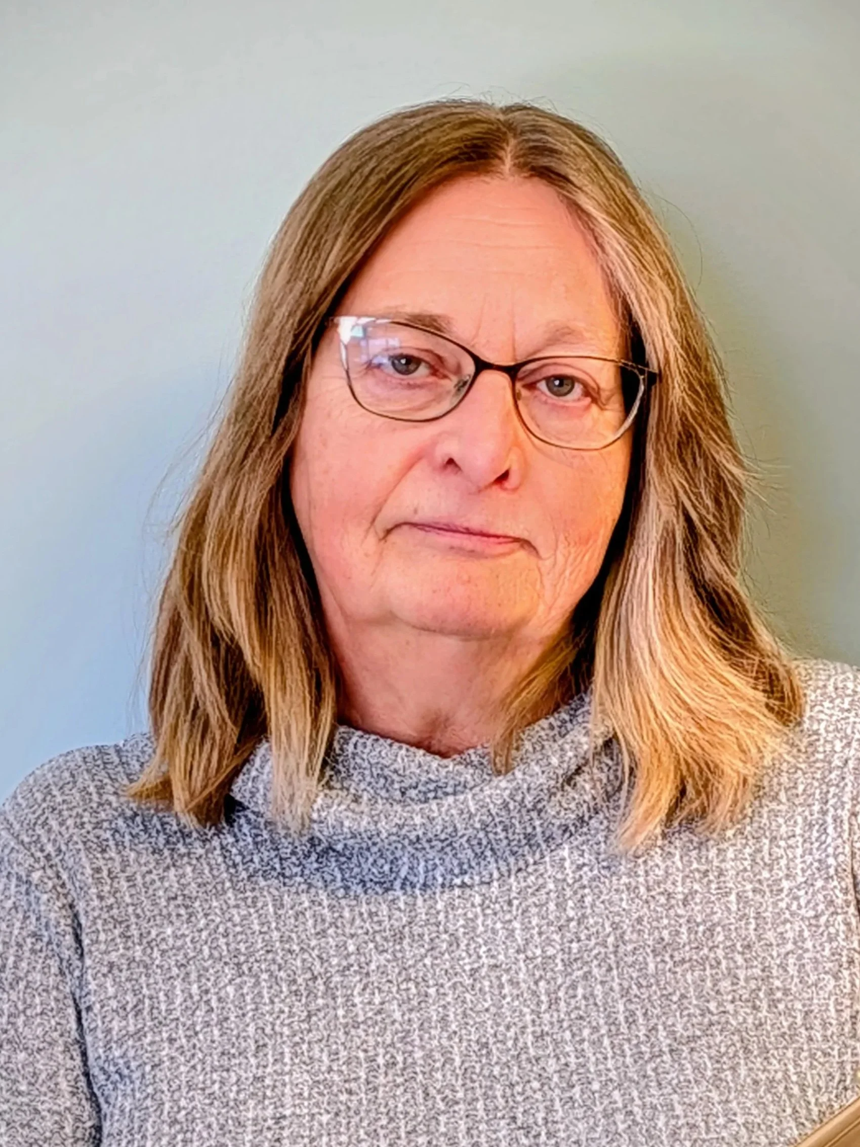 A woman with dark blond hair and glasses stands in front of a greyish blue wall. She is wearing a grey sweater and holding an open file.