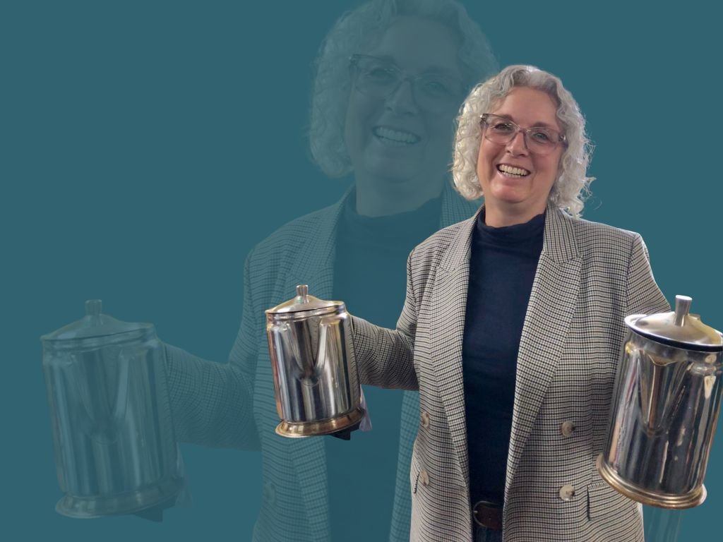 A smiling woman, wearing glasses and a plaid blazer over a dark top, is holding up two silver coffee pots.