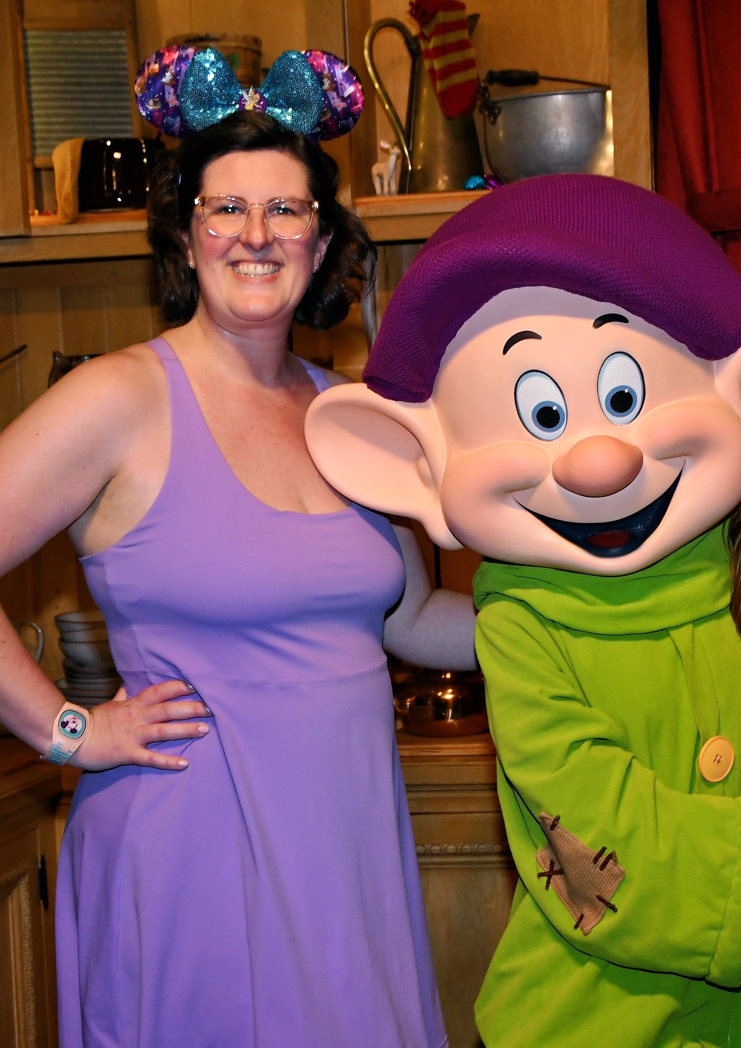 A woman wearing glasses, a purple dress, and Minnie Mouse ears headband, standing next to a character mascot of Dopey from Snow White. The woman is smiling and has her hand on her hip, while Dopey is smiling and dressed in a green outfit with a purple hat.
