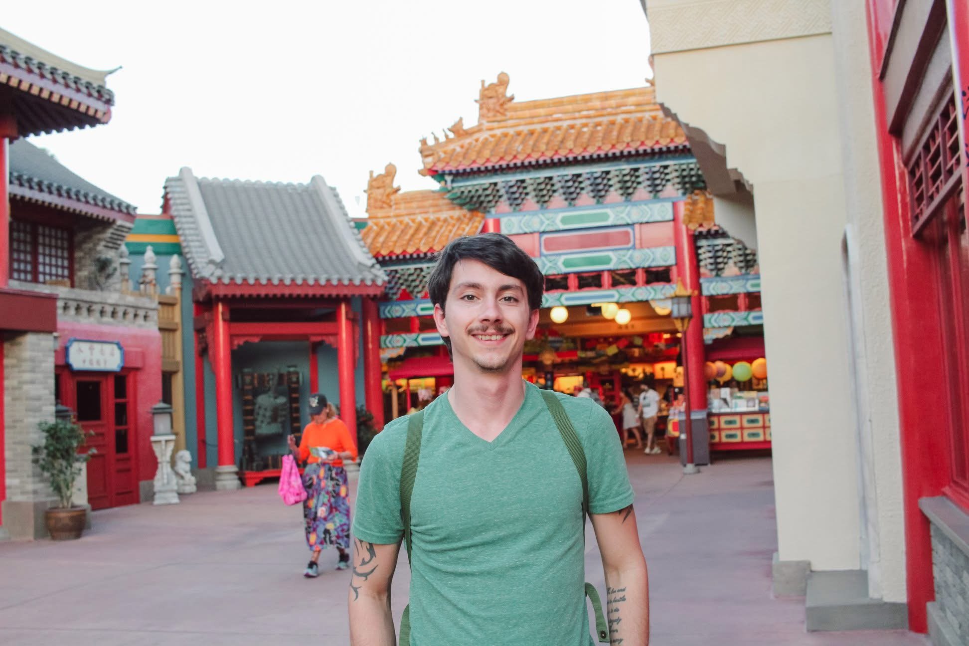 A young man with a green shirt and backpack standing in front of a colorful building with traditional Chinese architecture, including tiled roofs and red columns, at an amusement park or cultural site.