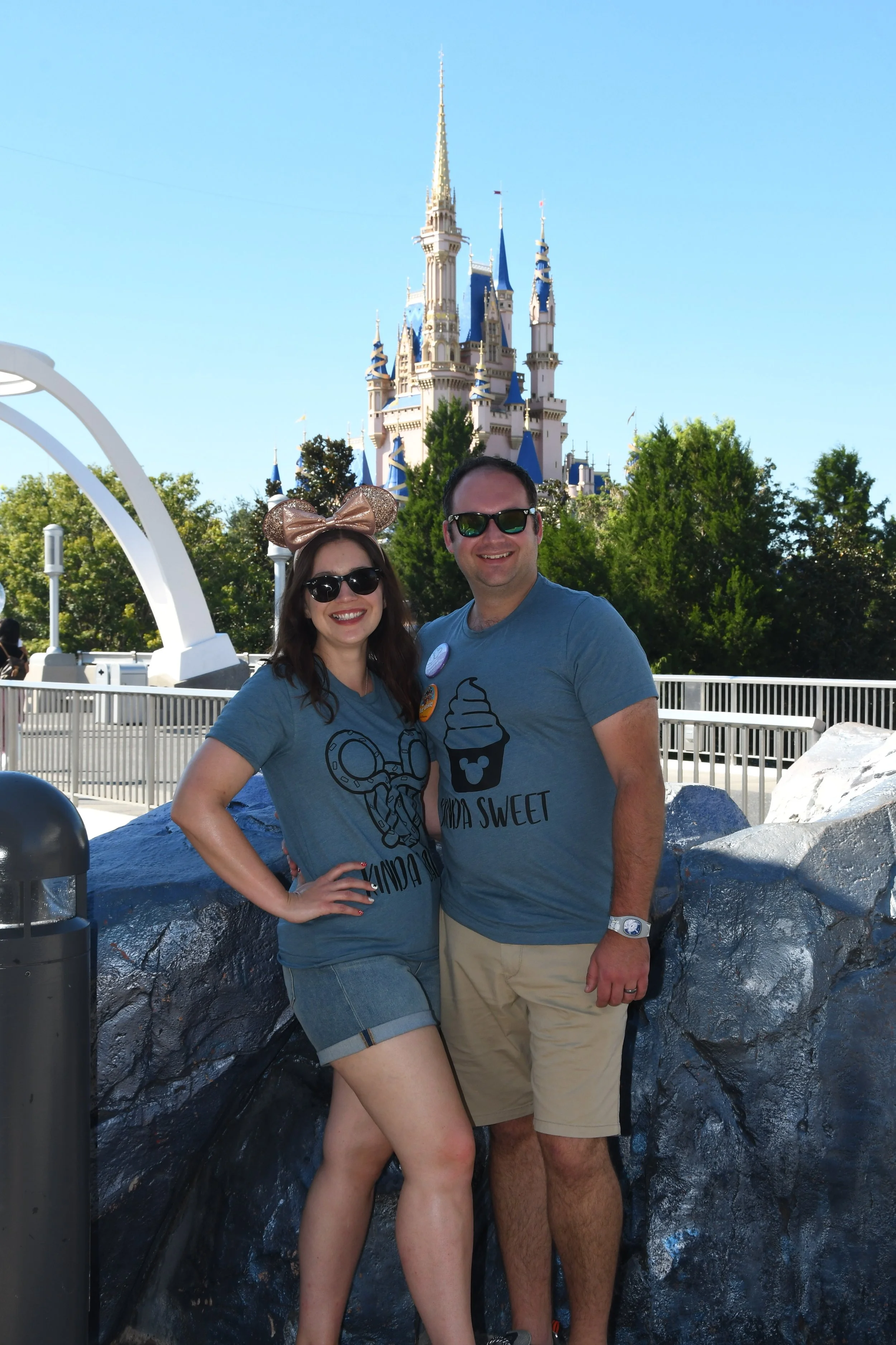 Young woman wearing leopard print Minnie Mouse ears headband, black sunglasses, black sleeveless top, khaki shorts, and a necklace, smiling outdoors.