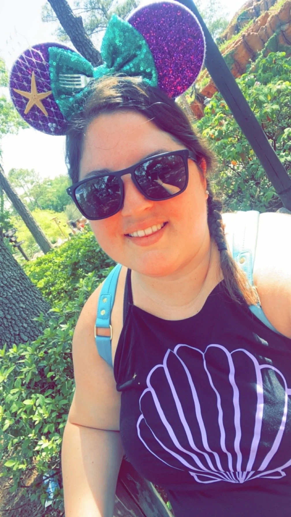 A woman with dark hair in a braid, wearing sunglasses and a black sleeveless top with a white shell outline, stands outdoors in a sunny environment with trees and greenery, souvenir Minnie Mouse ears with purple, teal, and pink glitter and starfish decorations on her head, and a light blue backpack strap visible.