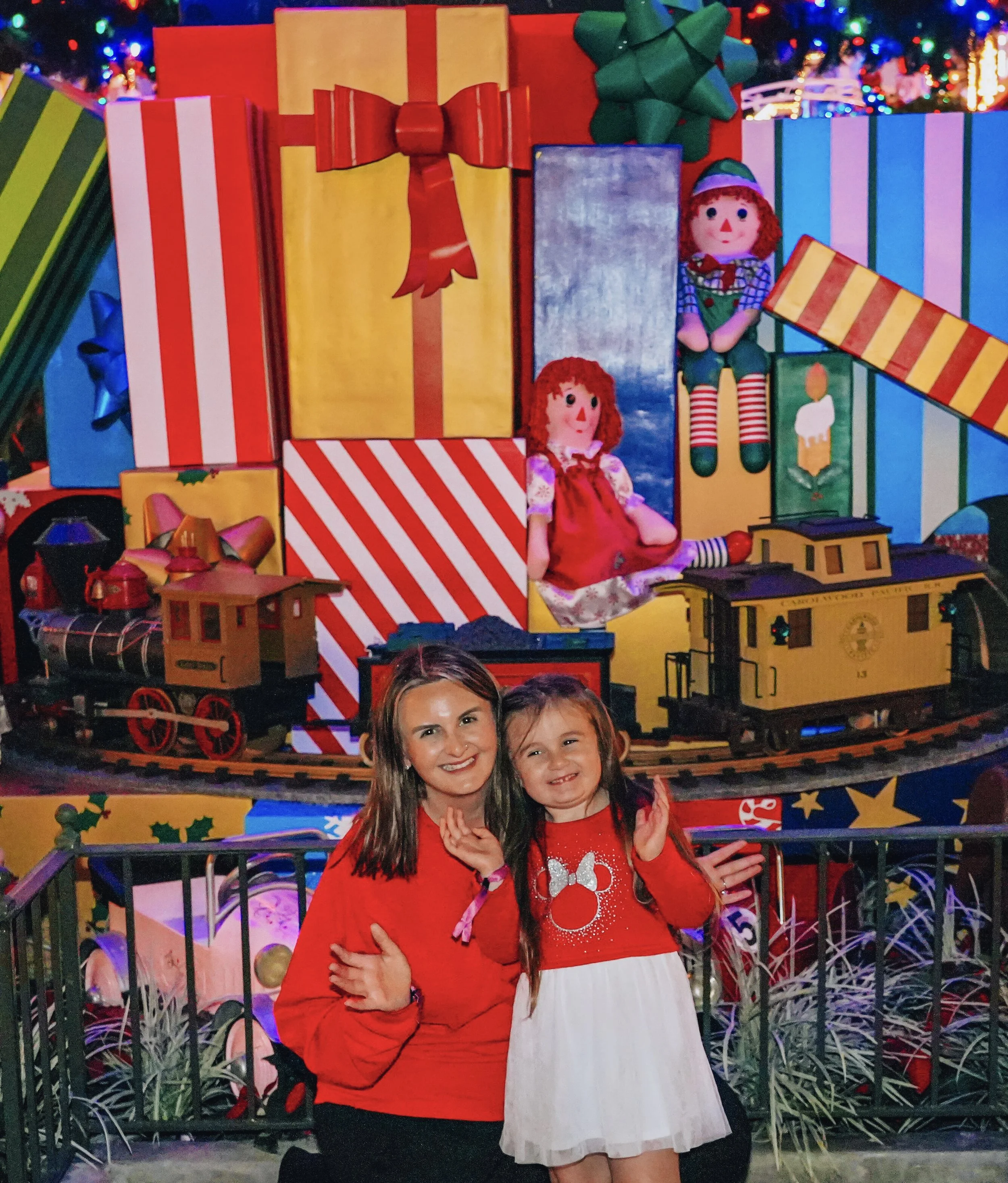 A woman and a young girl smiling and hugging in front of a festive Christmas display with large colorful gift boxes, dolls, toy train, and Christmas lights.