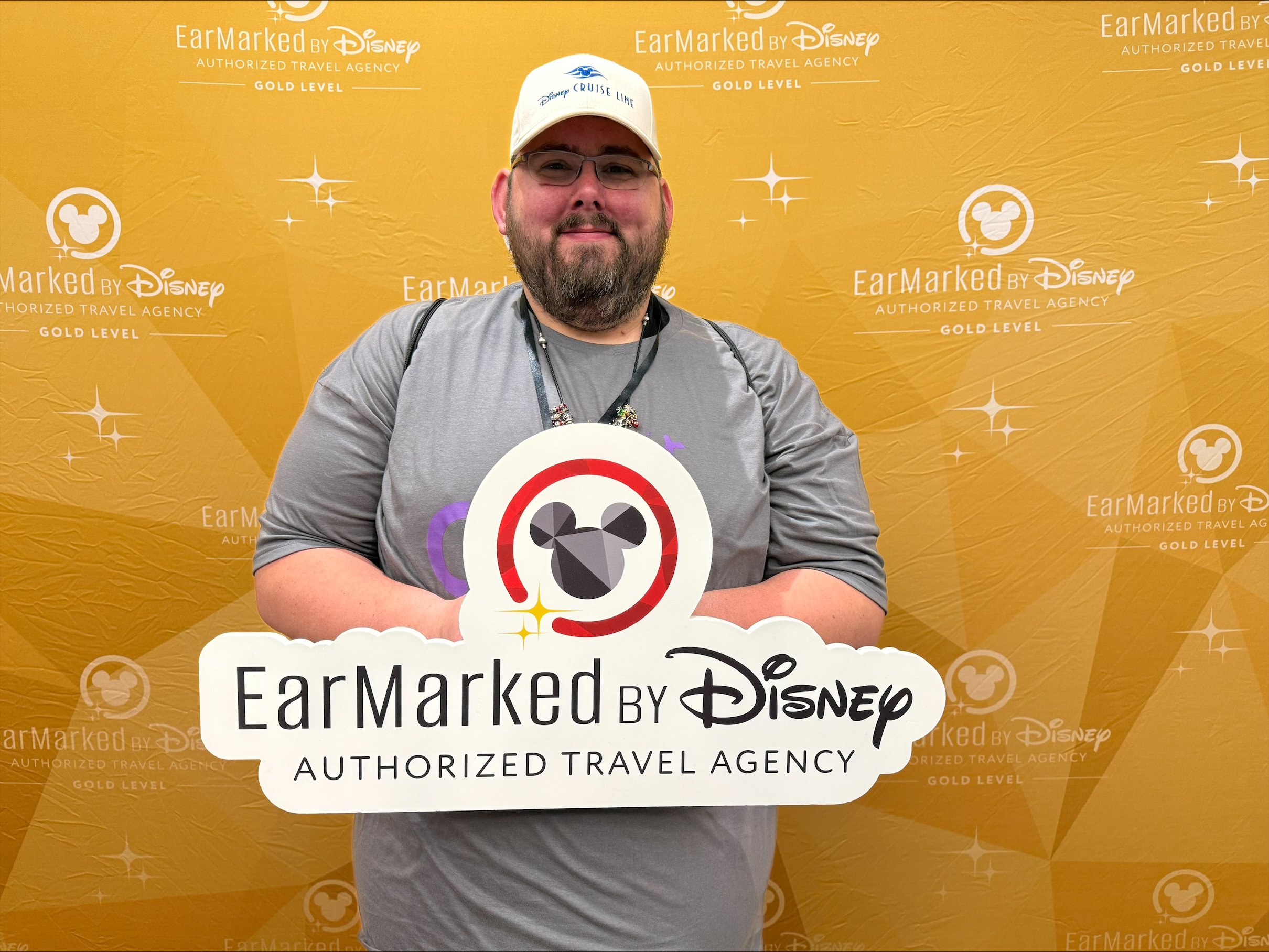 A man with glasses, a beard, wearing a blue patterned shirt and gold chain, standing in front of a painting of a cartoonish lion dressed as a historical figure.