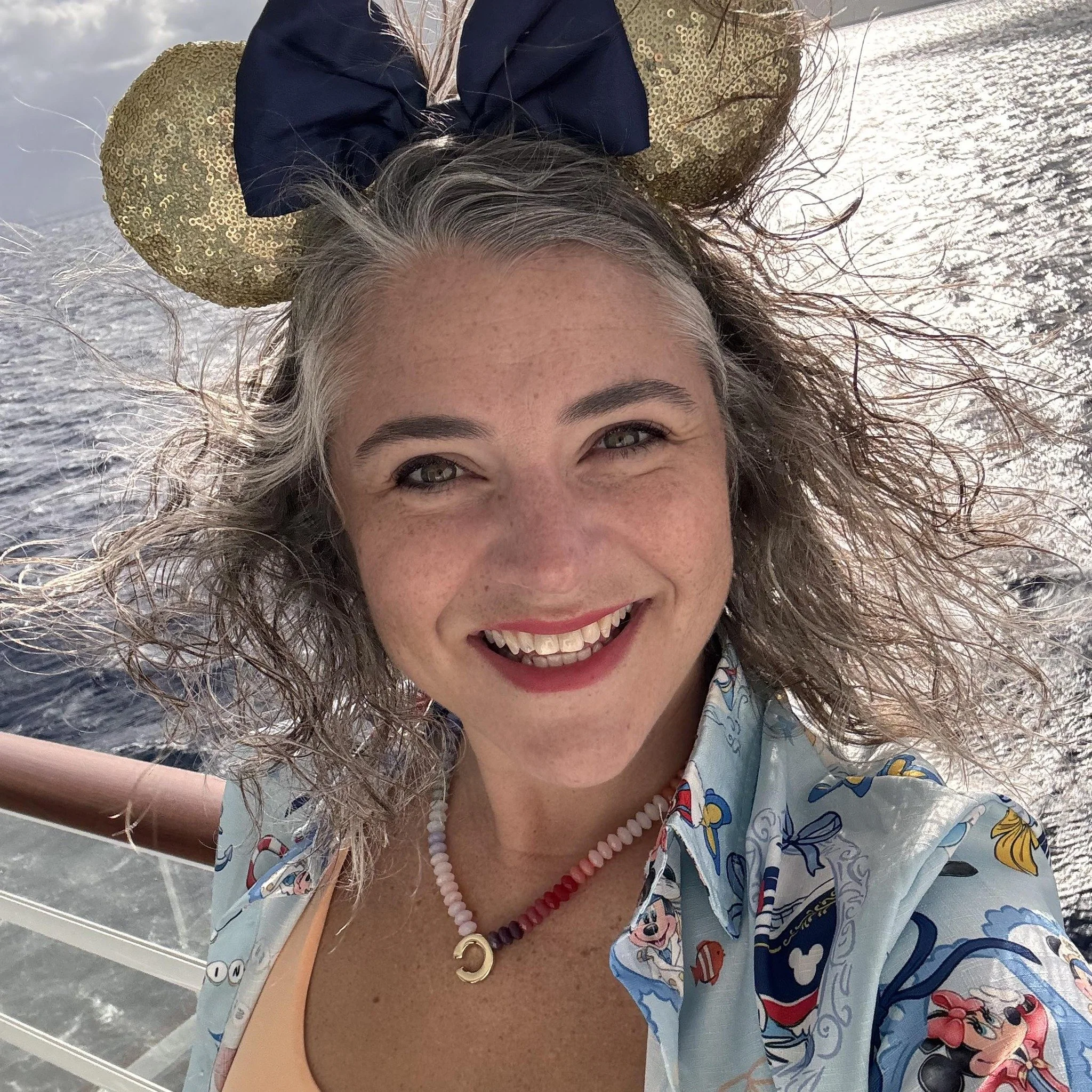 A smiling woman with curly gray hair, wearing a Disney-themed shirt and a Mickey Mouse ears headband, taking a selfie on a boat with water in the background.