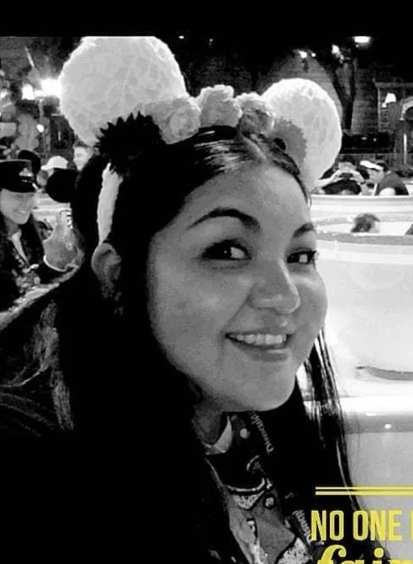 A woman with long dark hair wearing a headband with large, fluffy white pom-poms, smiling at the camera in a bustling indoor setting, possibly a restaurant or theme park.