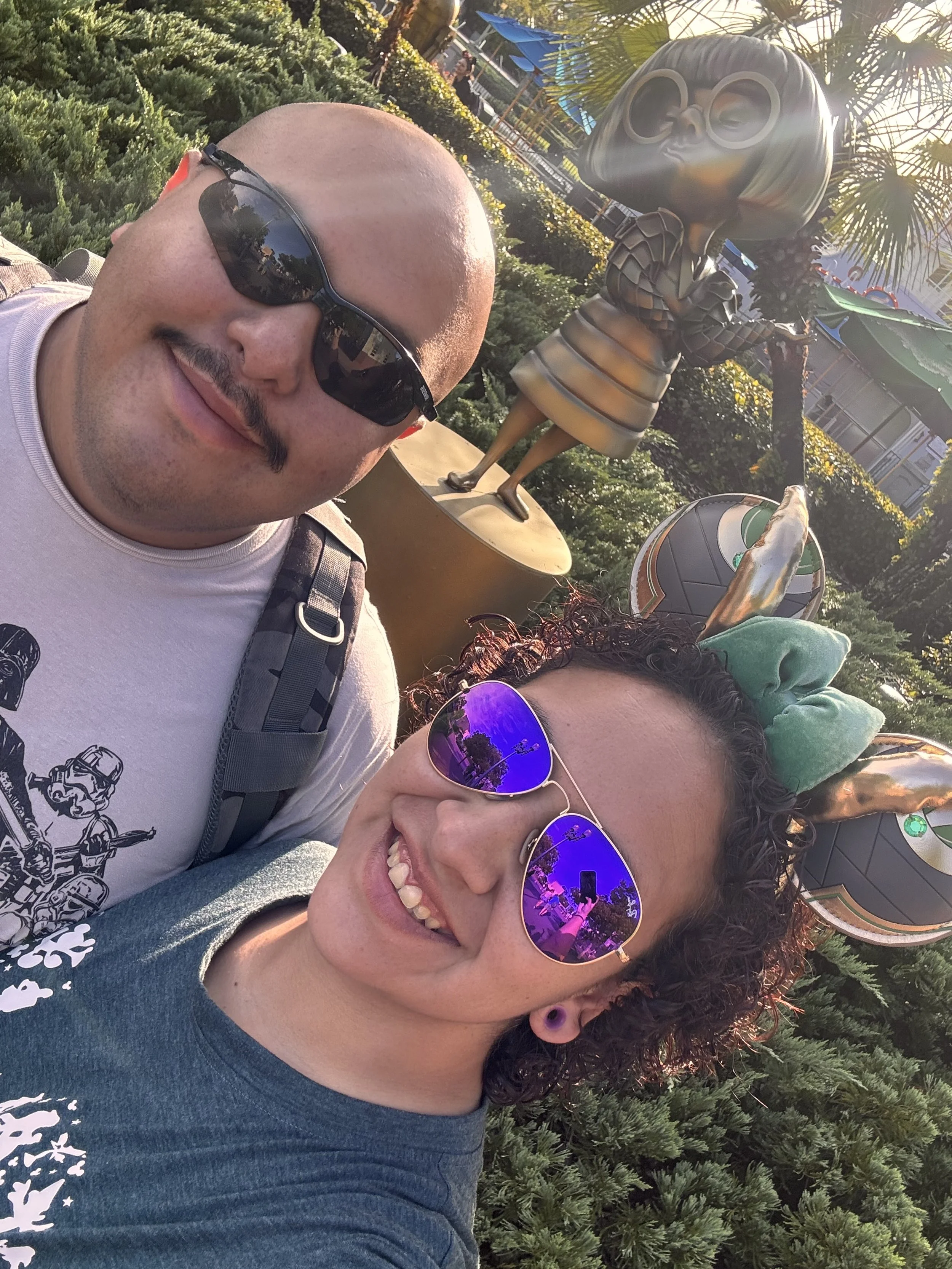 Two people taking a selfie outdoors, wearing sunglasses and smiling, with Disney's Epcot statue of a girl with glasses and a dress in the background, along with greenery and trees.