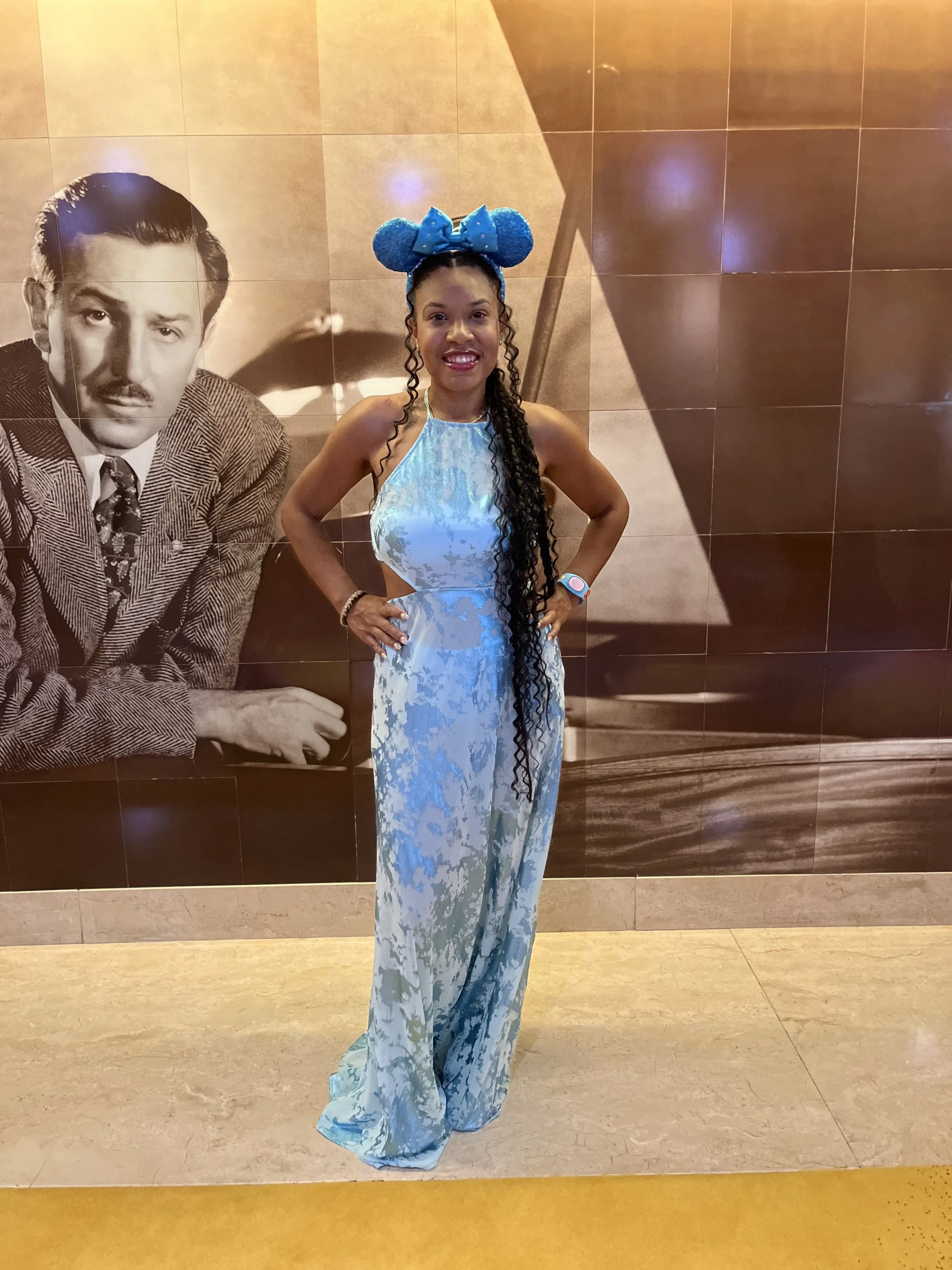 Woman in a blue patterned halter dress and matching Disney theme headband with ears, standing in front of large black and white photograph of a man in a suit.