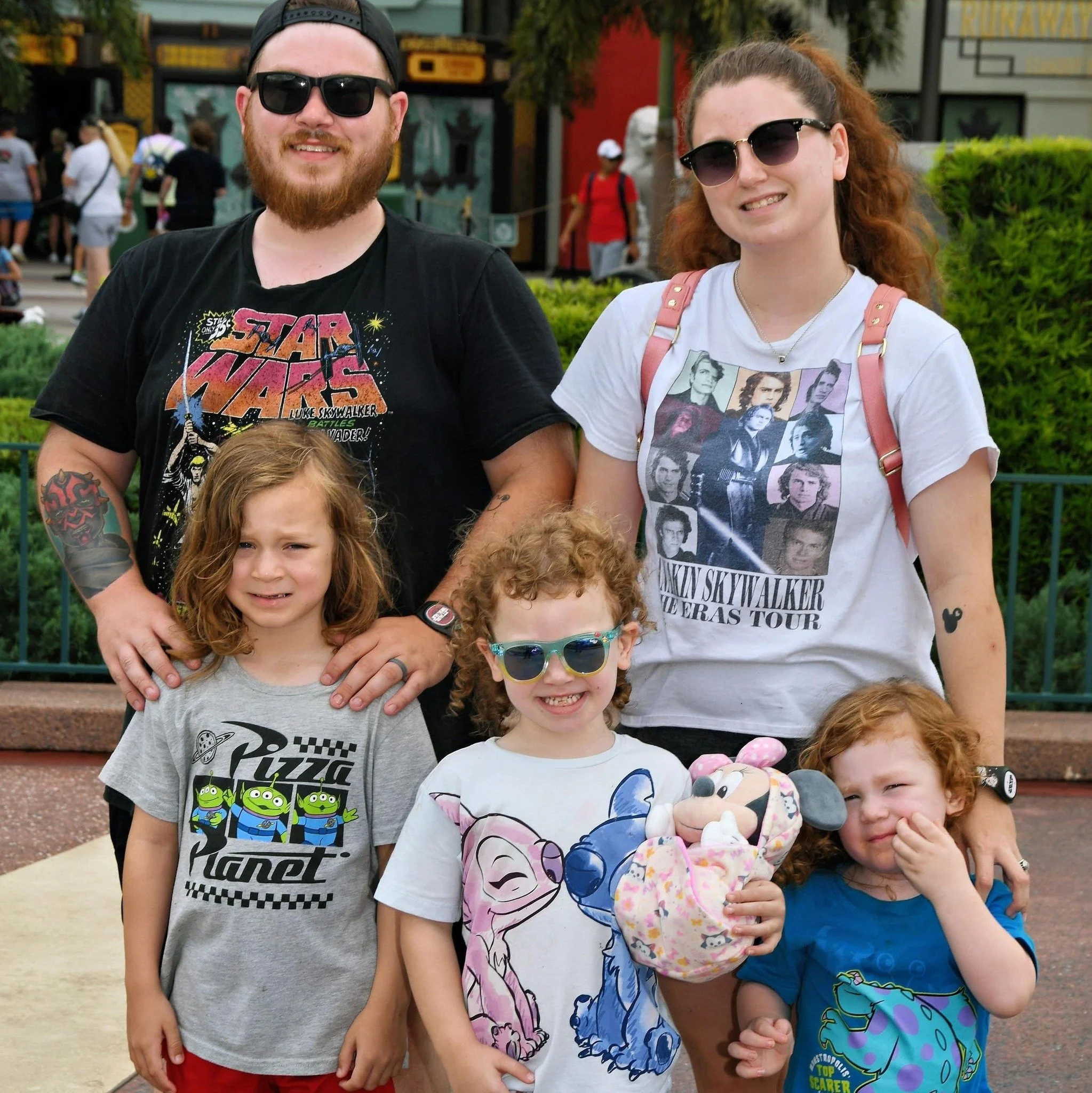 A family of six posing together at an amusement park, with two adults and four children, all smiling and wearing casual clothing and sunglasses.