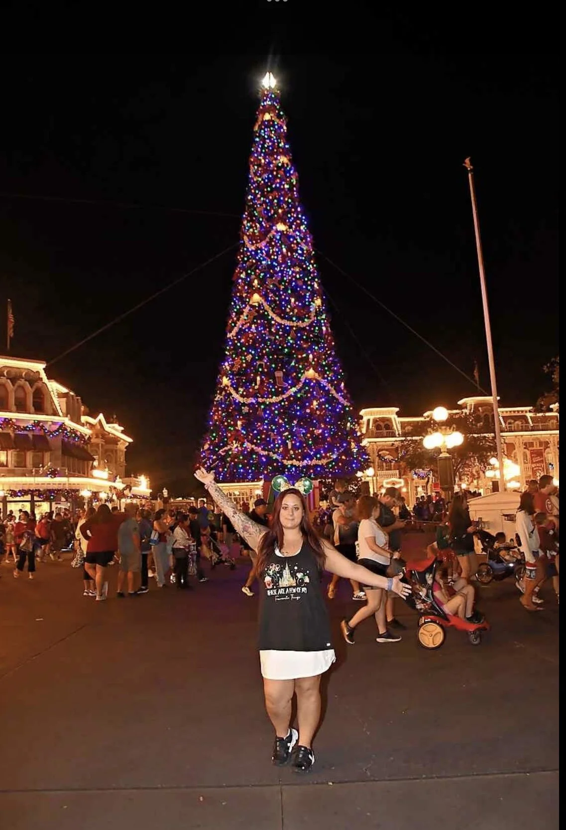 A woman standing with arms raised in front of a large, decorated Christmas tree at night, with many people and lit buildings in the background.