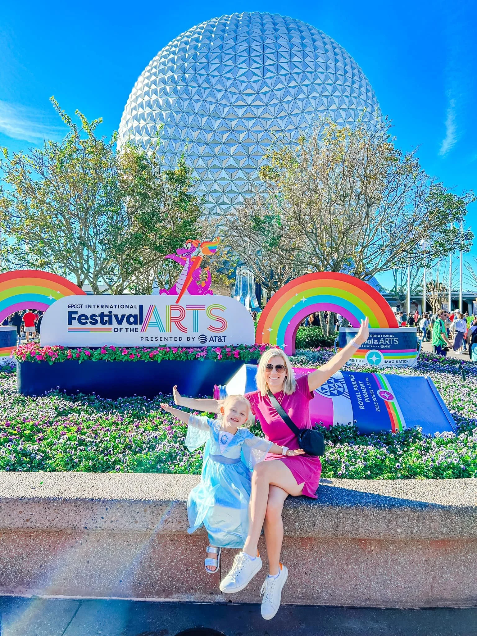 Two women sitting on a concrete ledge at EPCOT during the Festival of the Arts, with Spaceship Earth in the background, colorful rainbow decorations, and flowerbeds.