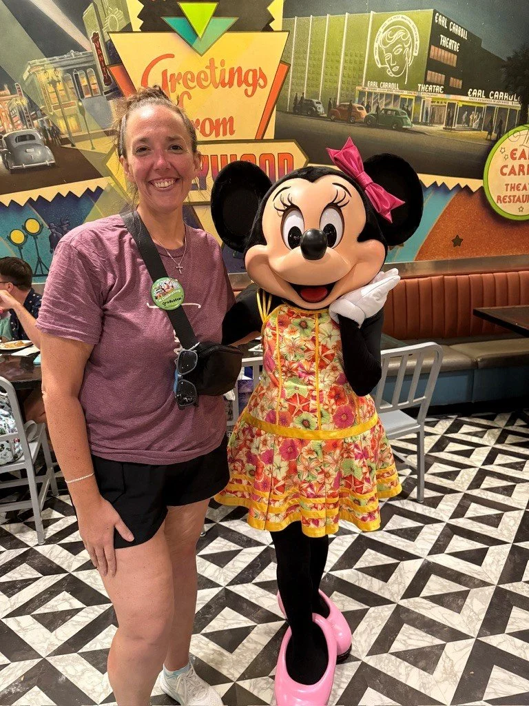 A woman smiling and standing next to Minnie Mouse, who is wearing a floral dress and pink shoes, inside a restaurant with vintage decor and colorful wall murals.