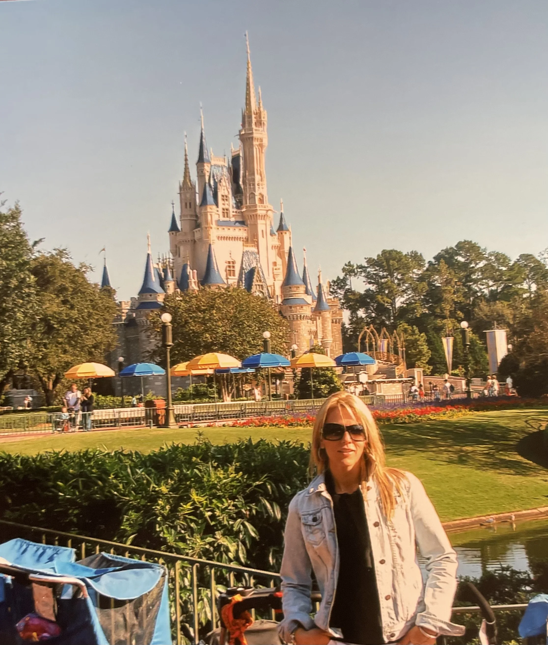 A woman with blonde hair and sunglasses standing in front of Sleeping Beauty Castle at Disneyland, with indoor garden and park visitors in the background.