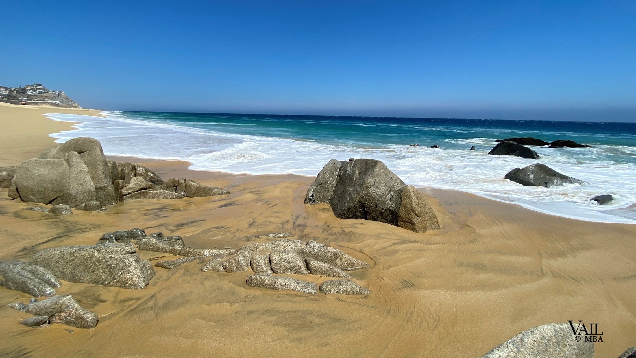Cabo San Lucas sandy beach with large rocks in the foreground, waves crashing onto the shore, and a clear blue sky. 