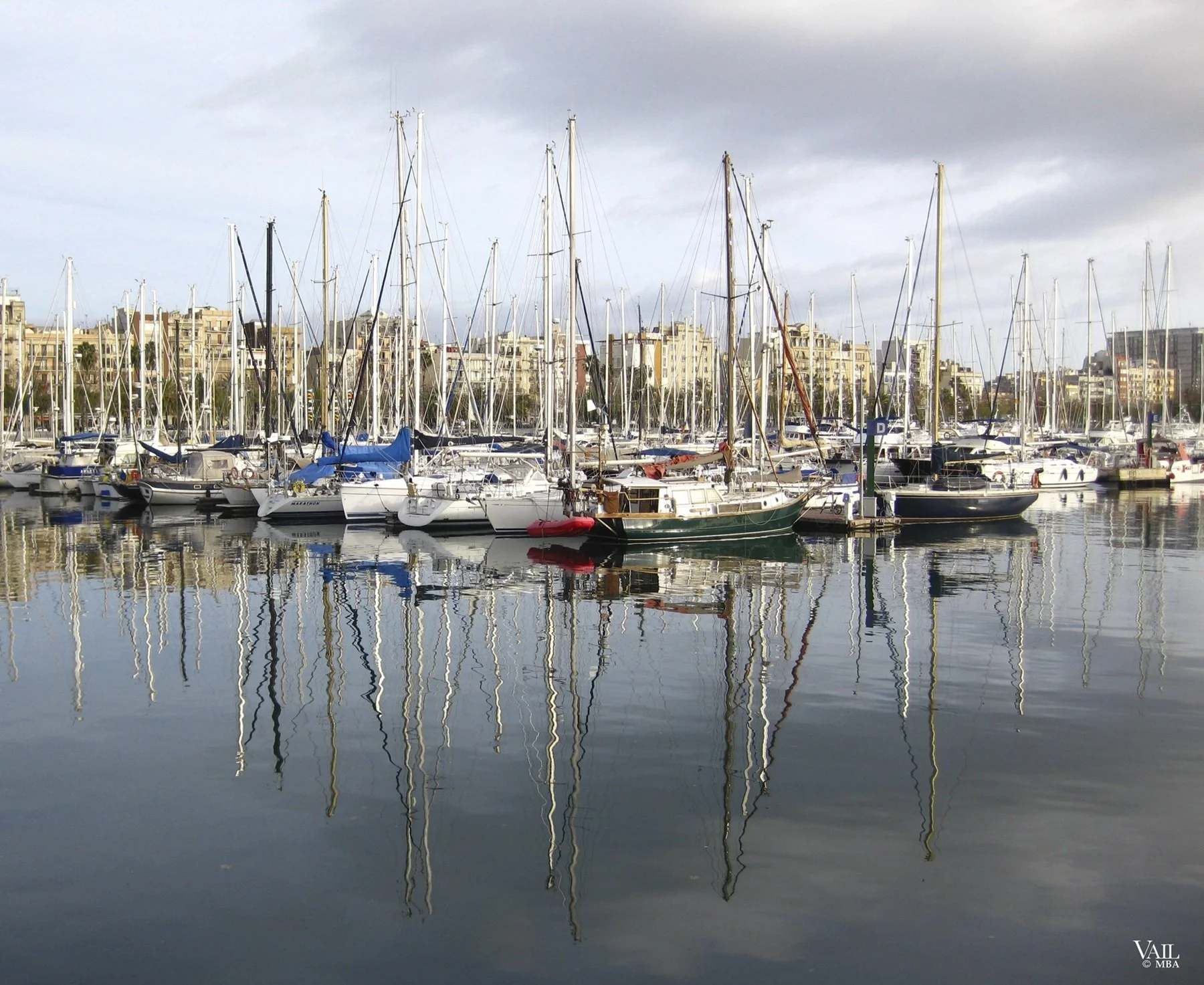 A marina in Barcelona, Spain with numerous sailboats docked in calm water, with tall masts and reflected image in the water, city buildings in the background, partly cloudy sky.