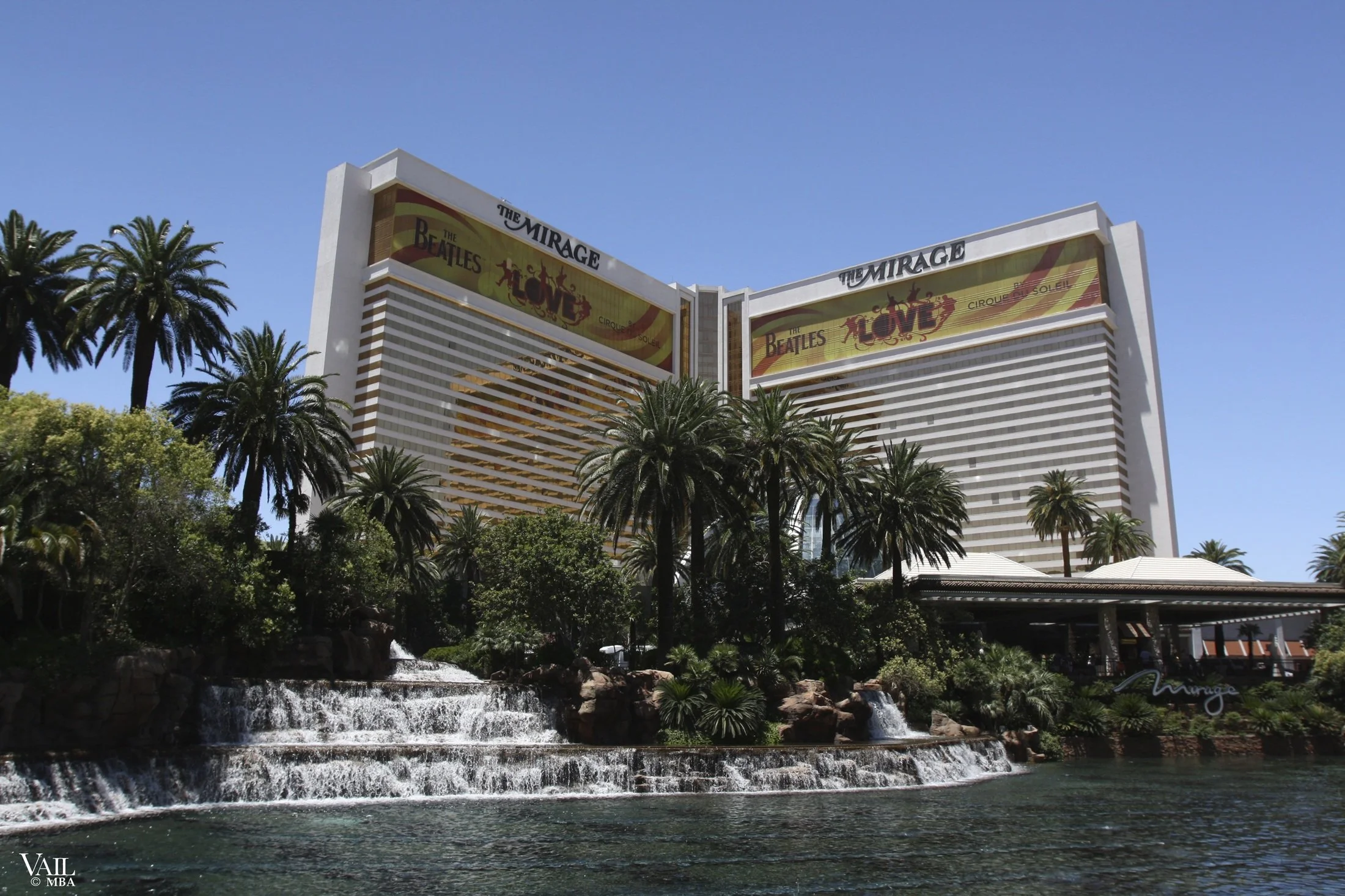 Daytime photo of the Mirage hotel in Las Vegas with a water feature and lush trees in the foreground.
