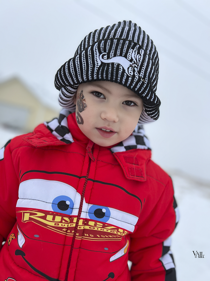 Young boy wearing a black and white striped beanie, red jacket with Lightning McQueen from Cars design, standing outdoors in a snowy environment.